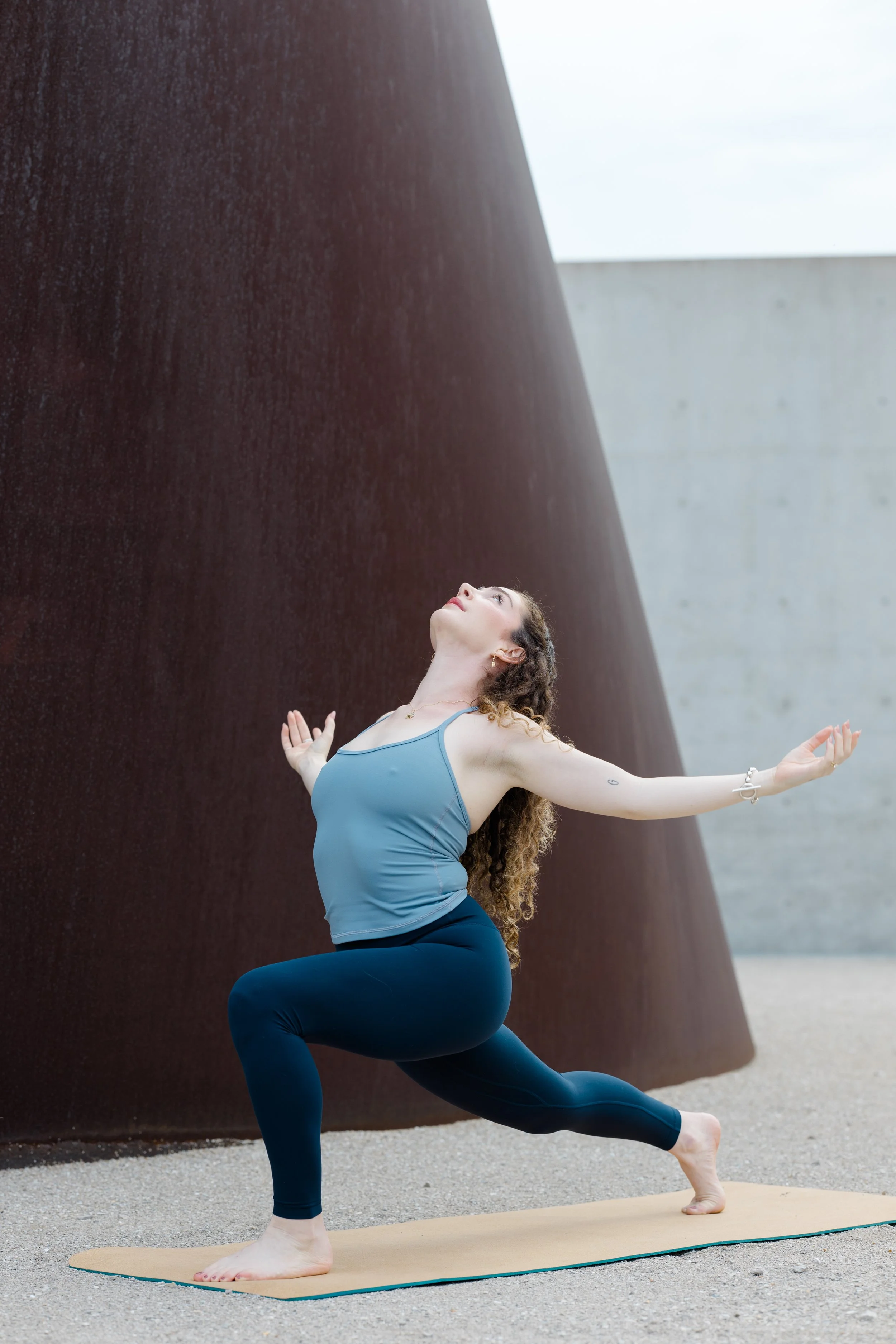 A woman practicing yoga outdoors on a yoga mat, in a pose with one knee bent and one leg extended back, arms raised and head tilted upward, with a large rust-colored structure behind her.