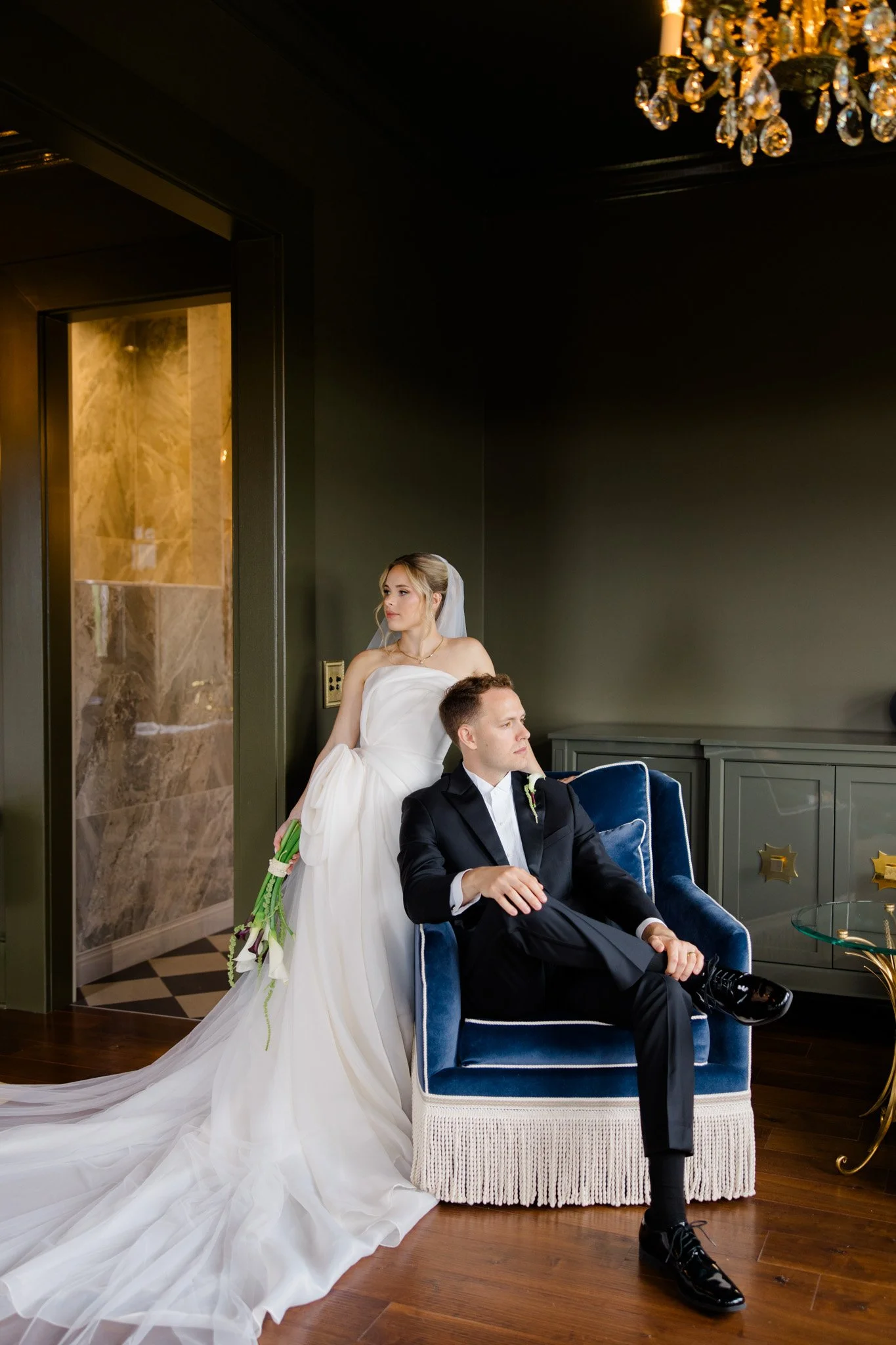 A bride in a white wedding gown and veil holding a bouquet stands behind a seated groom in a tuxedo, in an elegant room with a chandelier, dark walls, and a green cabinet.
