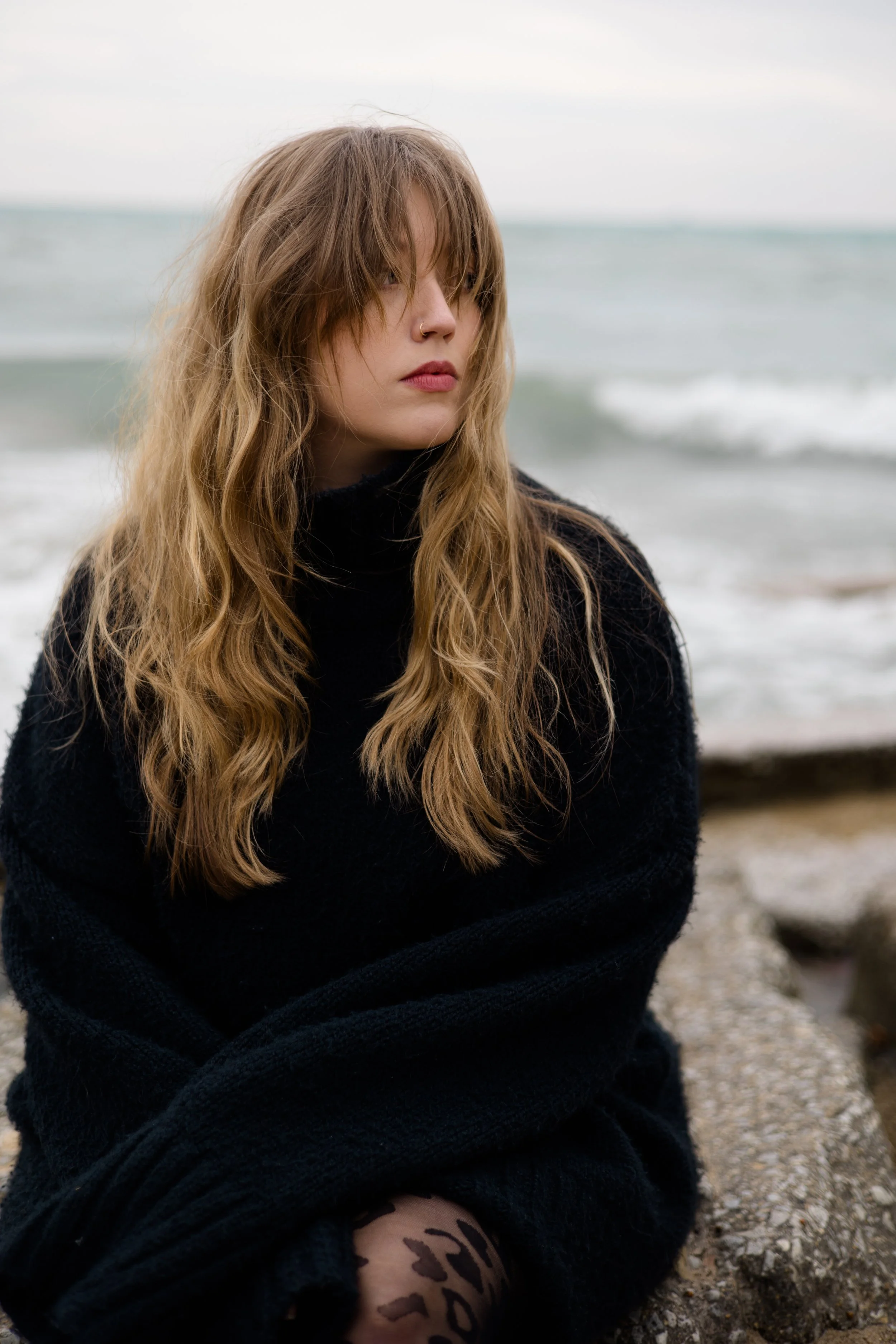 A young woman with long, wavy brown hair, wearing a black turtleneck, sitting by the beach with ocean waves in the background.