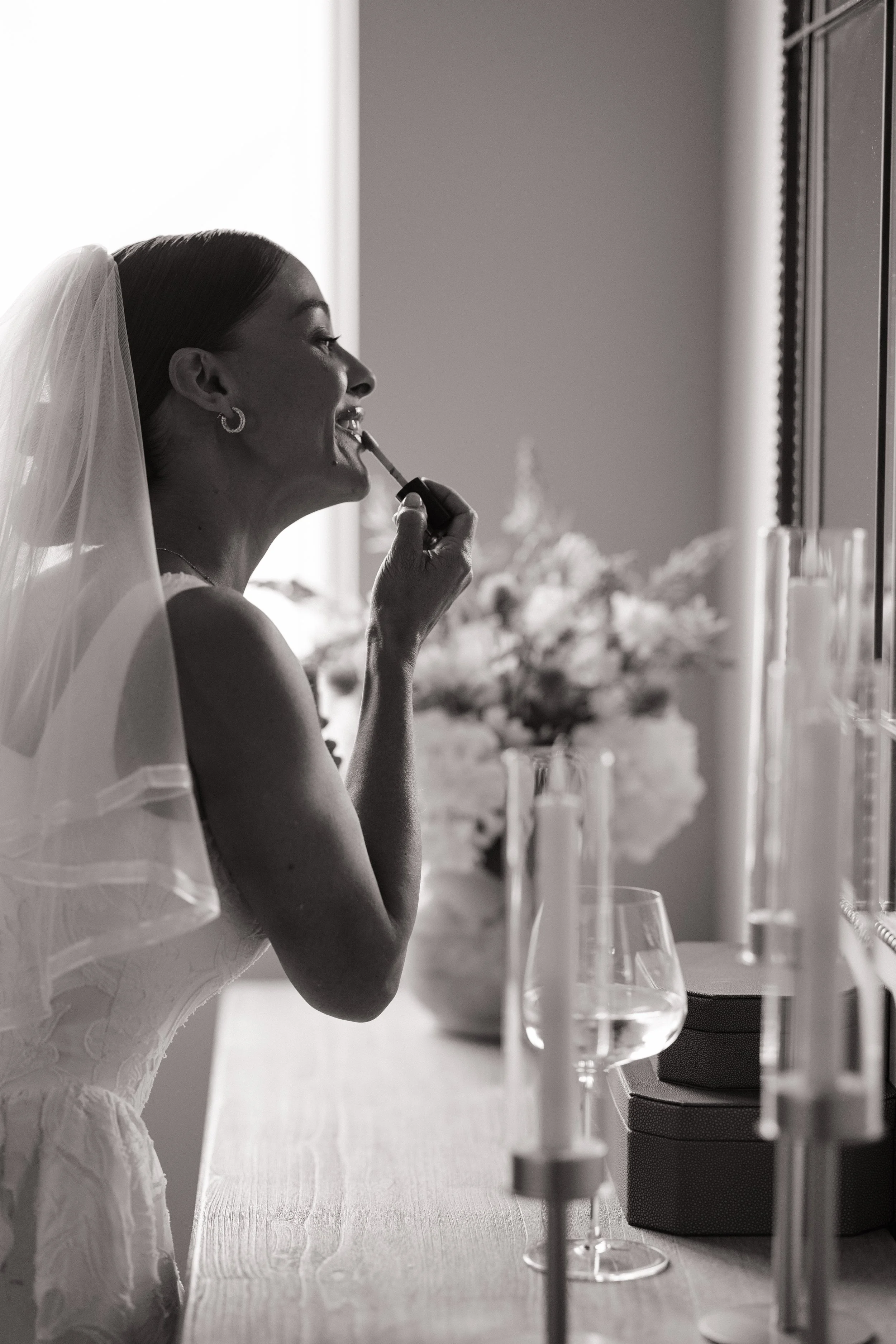 A woman in a wedding dress applies lipstick while sitting at a table with wine glasses and floral centerpiece.