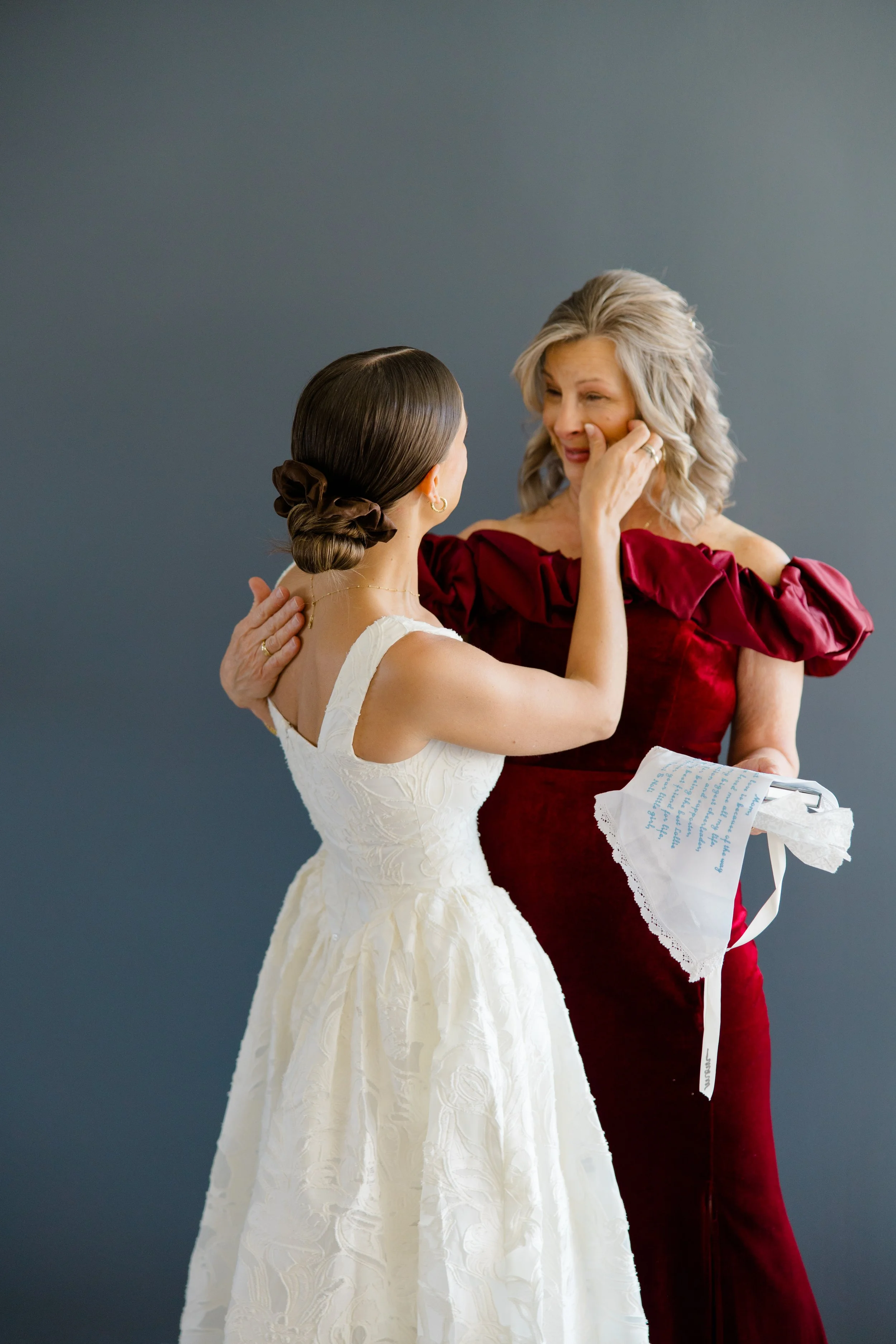 A woman in a white wedding dress touches the face of an older woman in a red dress, who is holding a piece of fabric with handwritten text. They are standing against a plain gray background.