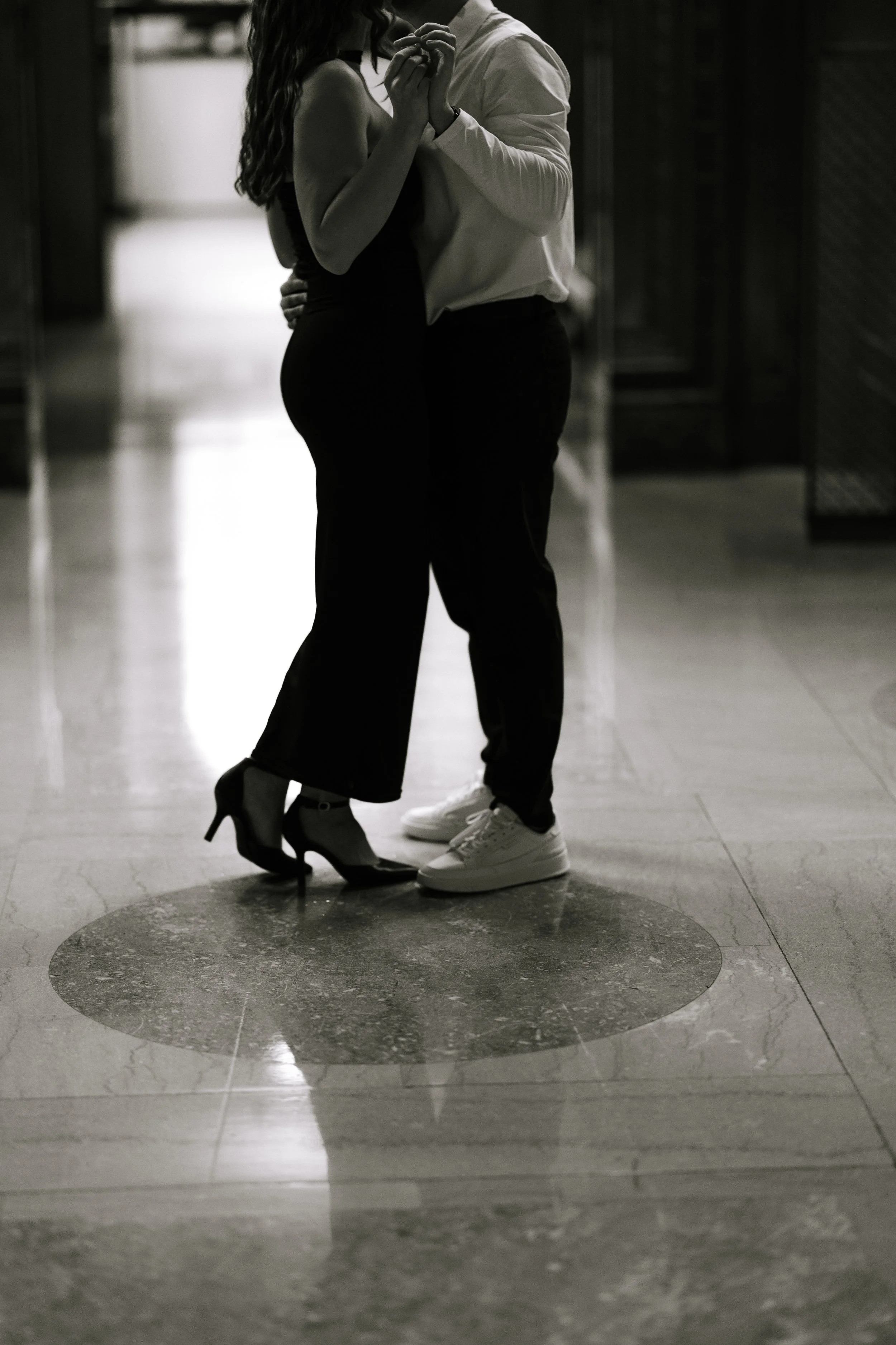 A black and white photo of a couple dancing closely, with their hands intertwined. The woman is wearing high heels and a sleeveless dress, and the man is wearing a long-sleeved shirt and sneakers. They are standing on a circular pattern on the floor,