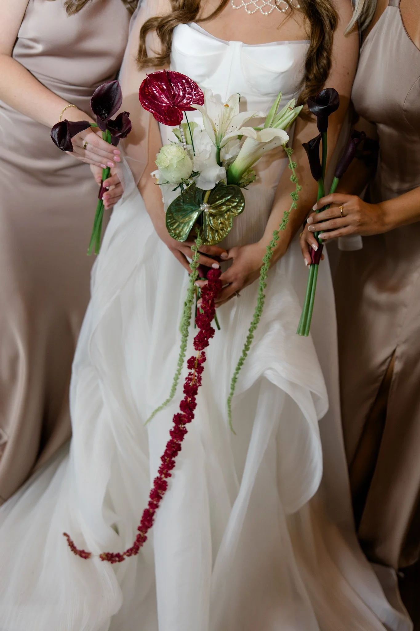 A woman in a white wedding dress holding a bouquet of white lilies, with her hands visible. Two women in beige dresses are holding long, dark-colored calla lilies on either side of her.