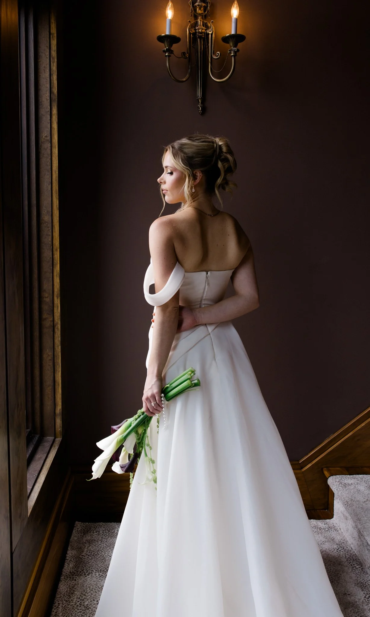 A woman in a wedding dress holding a bouquet of calla lilies, standing indoors near a dark wall and a wall sconce with two candles.