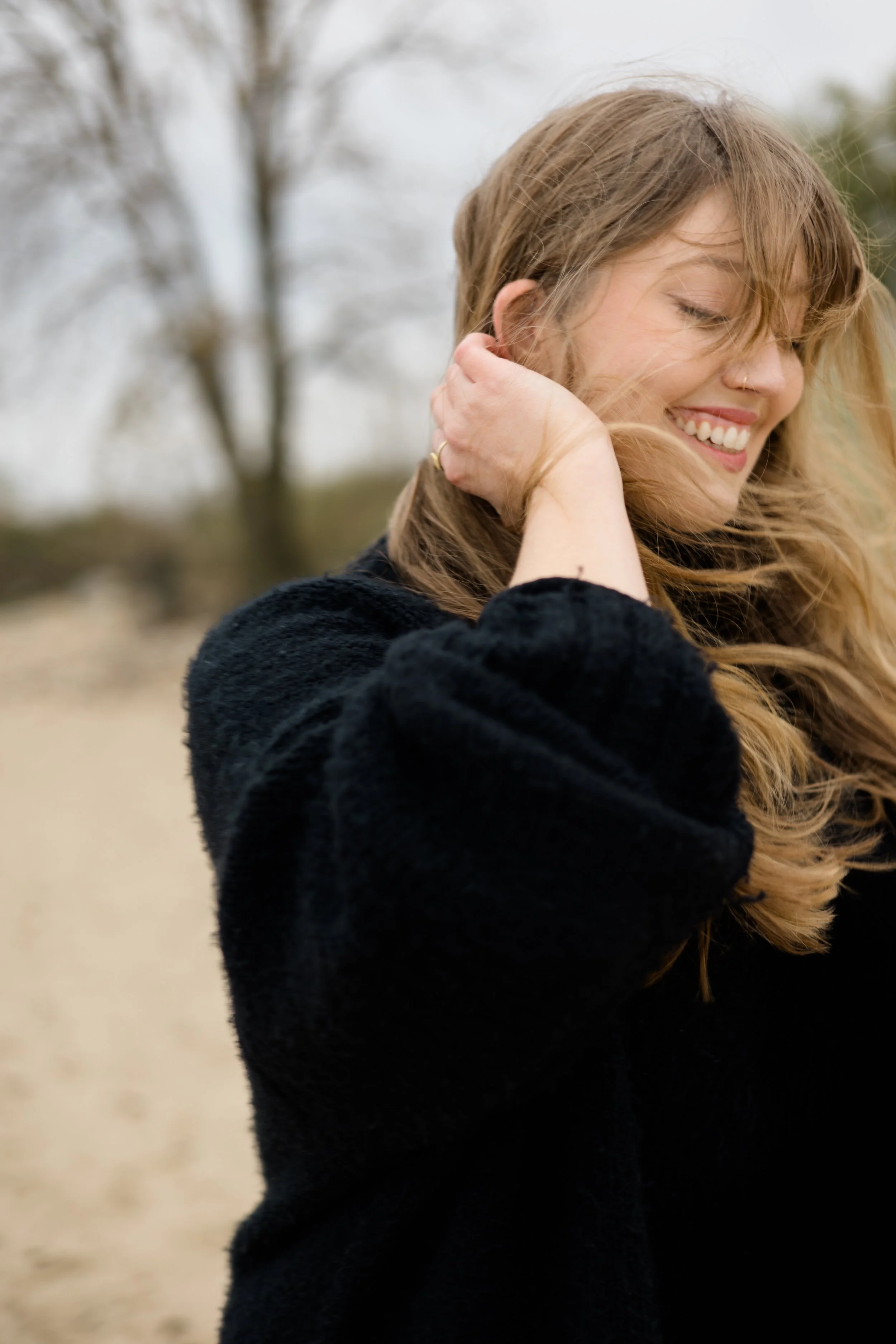 A woman with long, wavy hair smiling and touching her hair outdoors against a blurred background of trees.