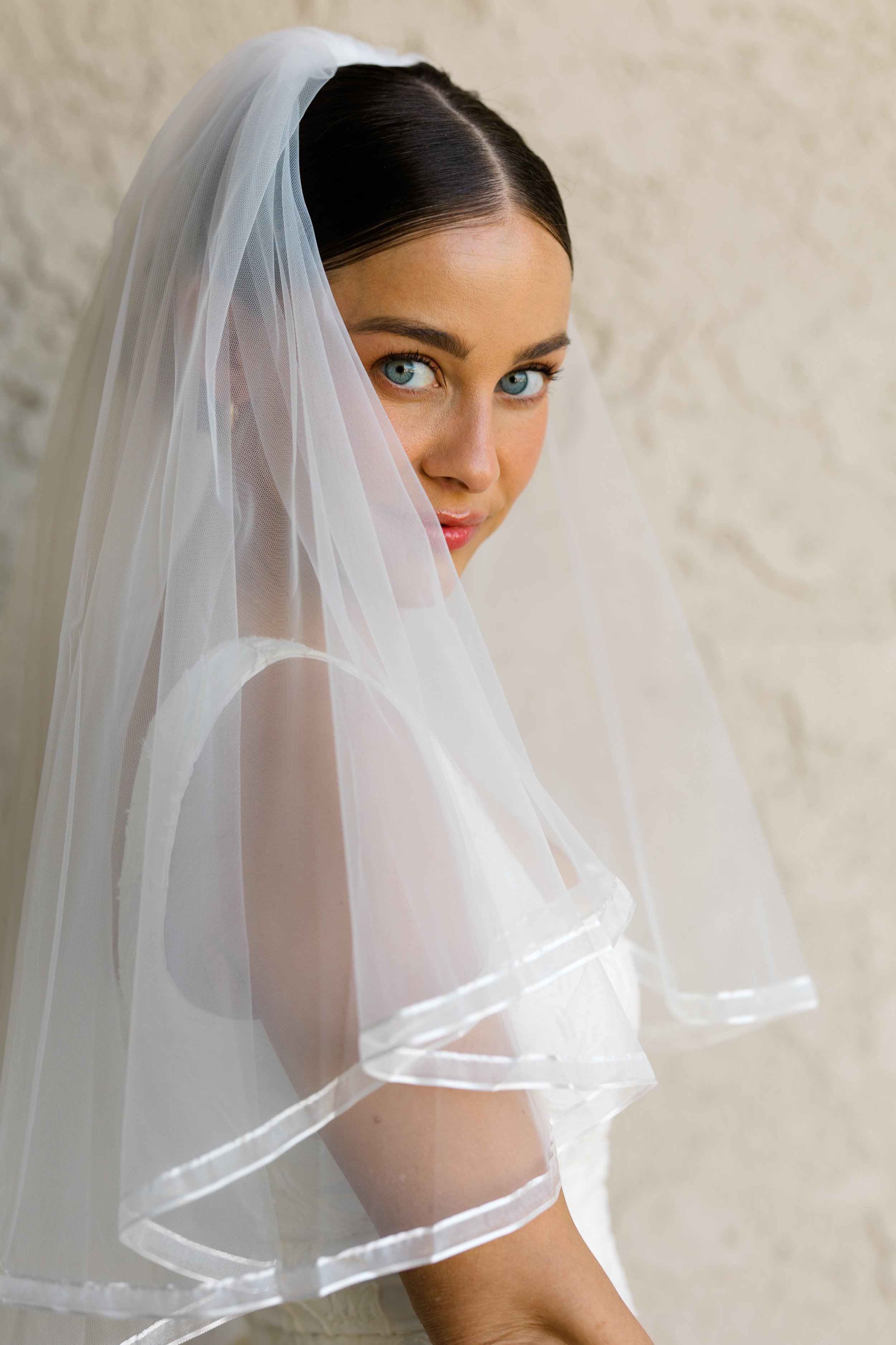 Close-up of a woman with blue eyes and dark hair, wearing a white veil with sheer fabric and satin trim, standing against a textured beige wall.