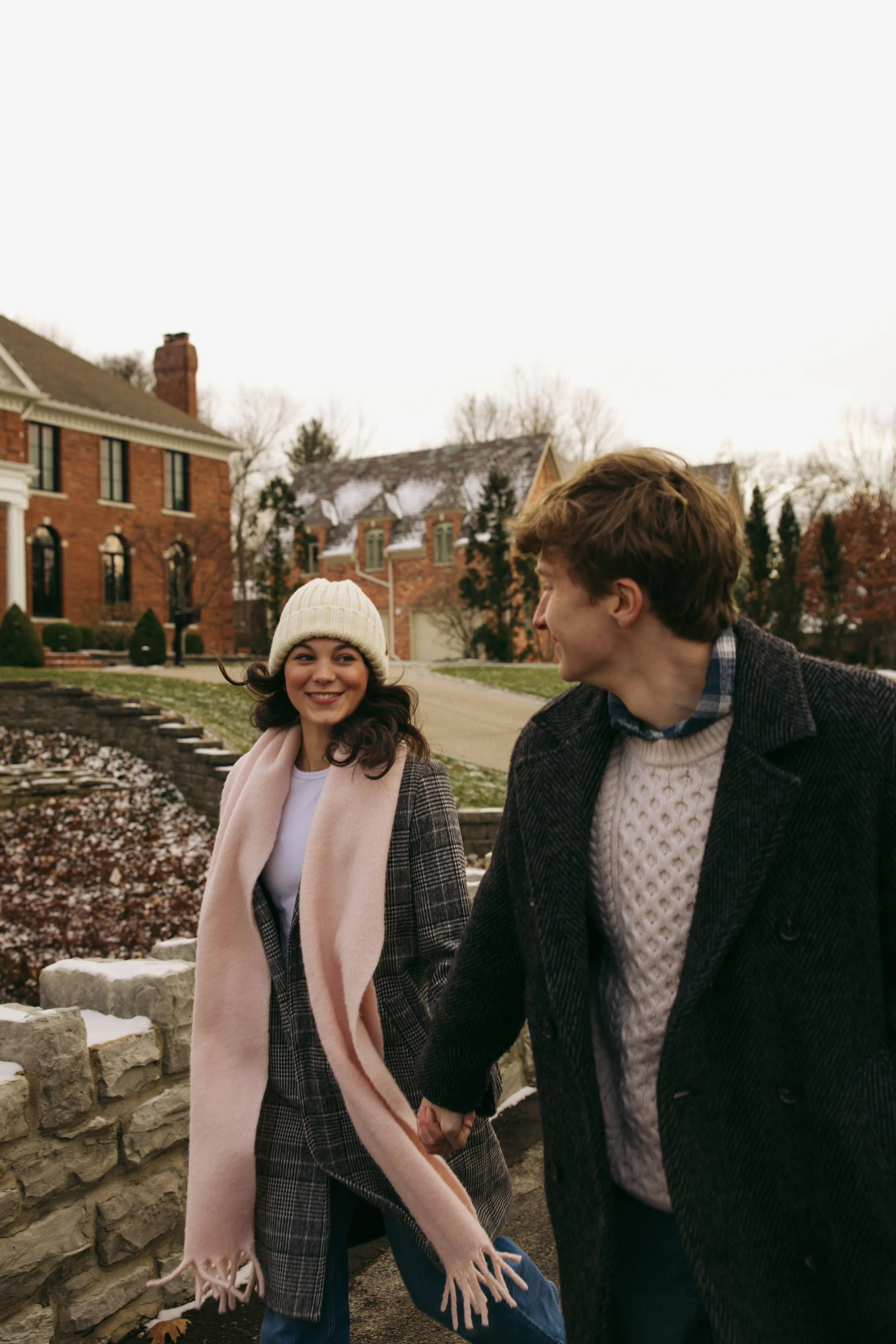 A young woman wearing a beige knit hat, plaid coat, and pink scarf smiling at a young man with brown hair, wearing a dark coat and white sweater, as they walk hand in hand on a sidewalk in a suburban neighborhood during fall or winter.