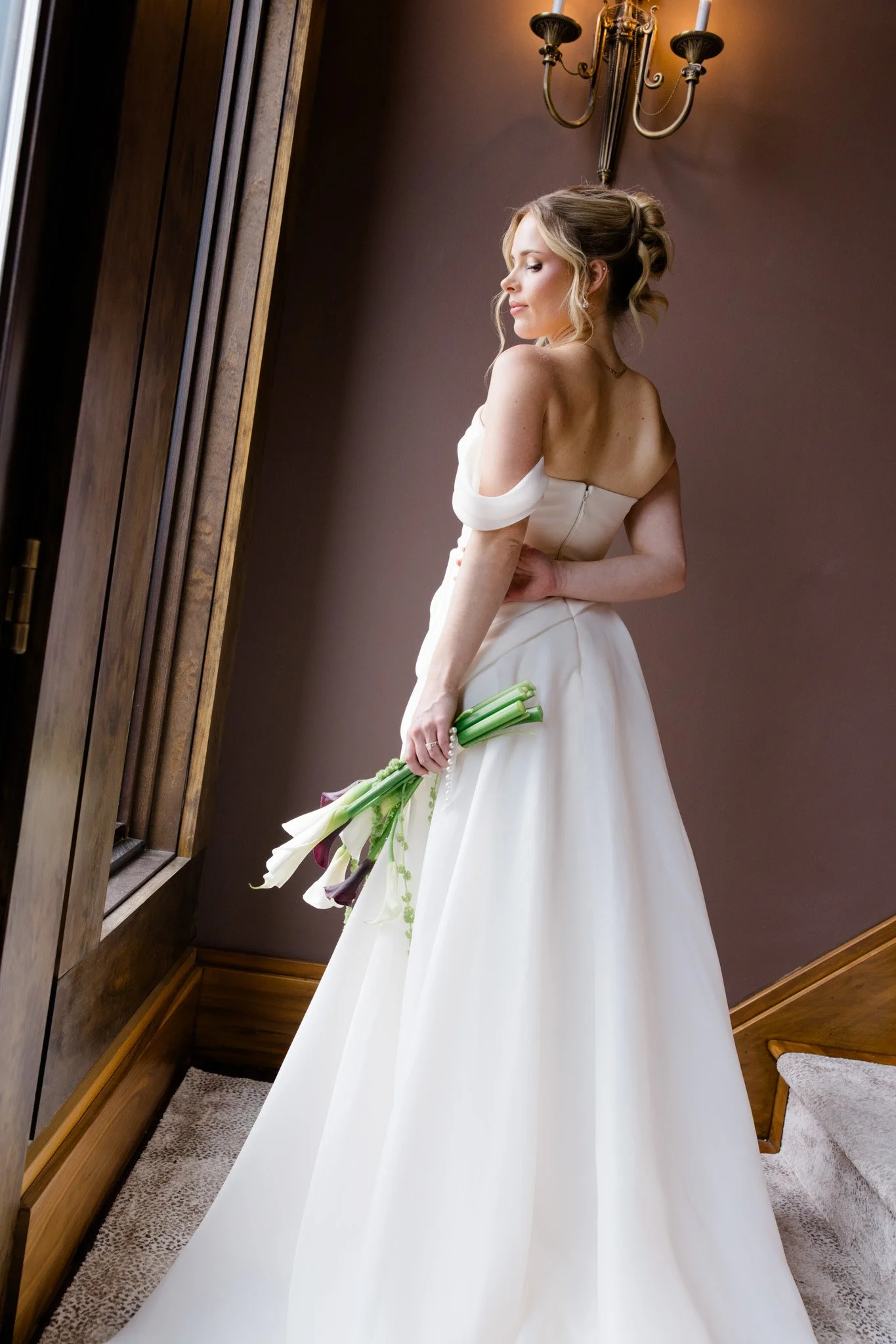 A bride in a white wedding gown holding a bouquet of calla lilies, standing next to a window with wooden trim in a room with brown walls and a chandelier lighting fixture.