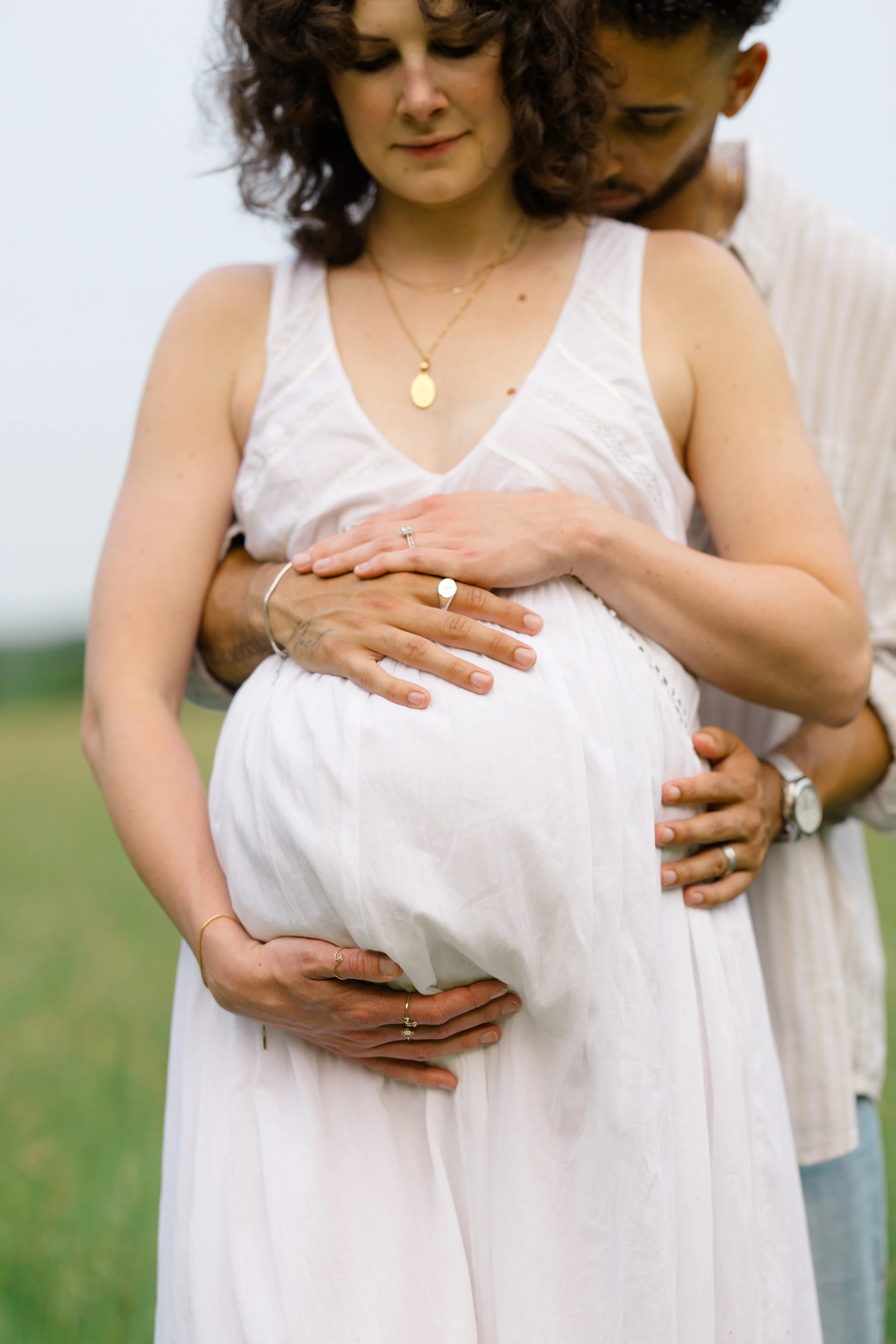 A pregnant woman wearing a white dress with her hands resting on her belly, being embraced from behind by a partner during daytime outdoors.