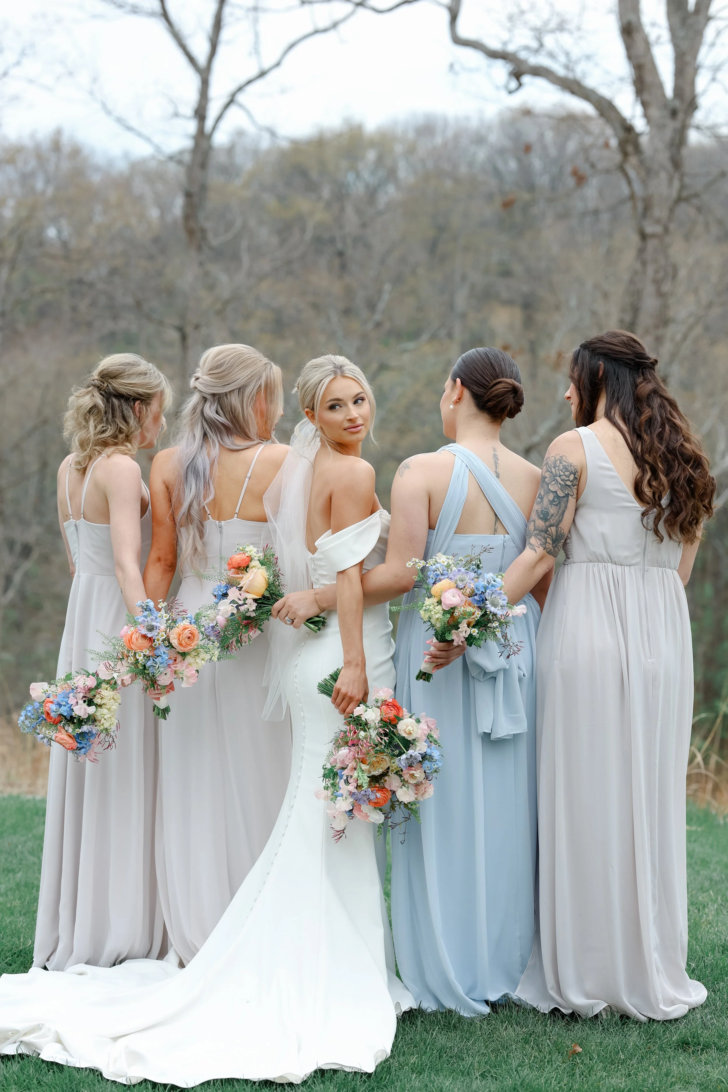 A bride standing in the middle of five bridesmaids outdoors with trees in the background, all holding bouquets of flowers, dressed in pastel-colored dresses, with the bride in a white gown and the bridesmaids in shades of blue and gray.