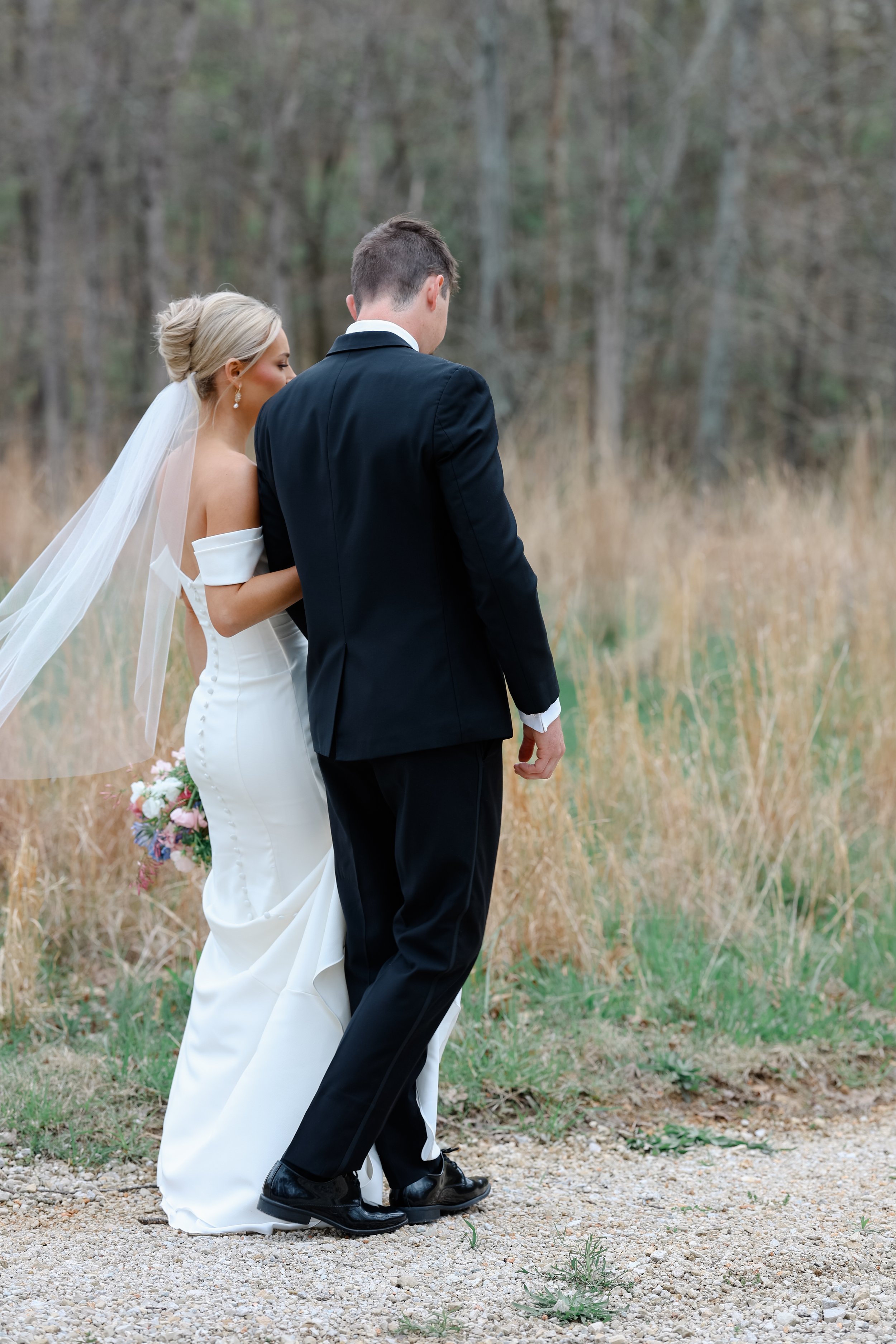 A bride and groom walking together outdoors in a natural setting, with the bride holding a bouquet of flowers and the bride wearing a white wedding dress with a veil, the groom in a black suit, and trees in the background.