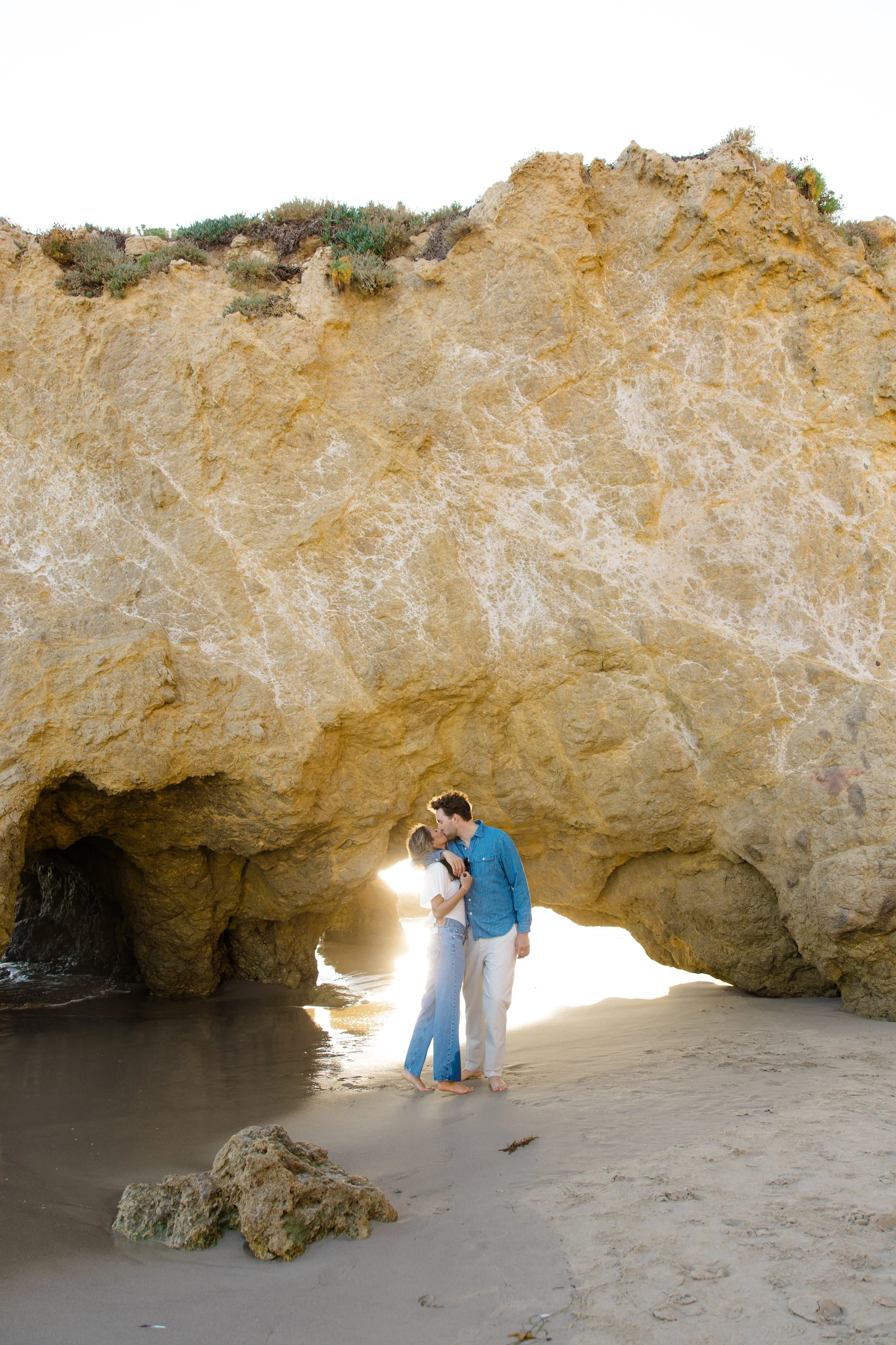 A couple in love sharing a kiss on a sandy beach at sunset, standing beneath a large rock formation with a cave-like opening, surrounded by rocks and water.