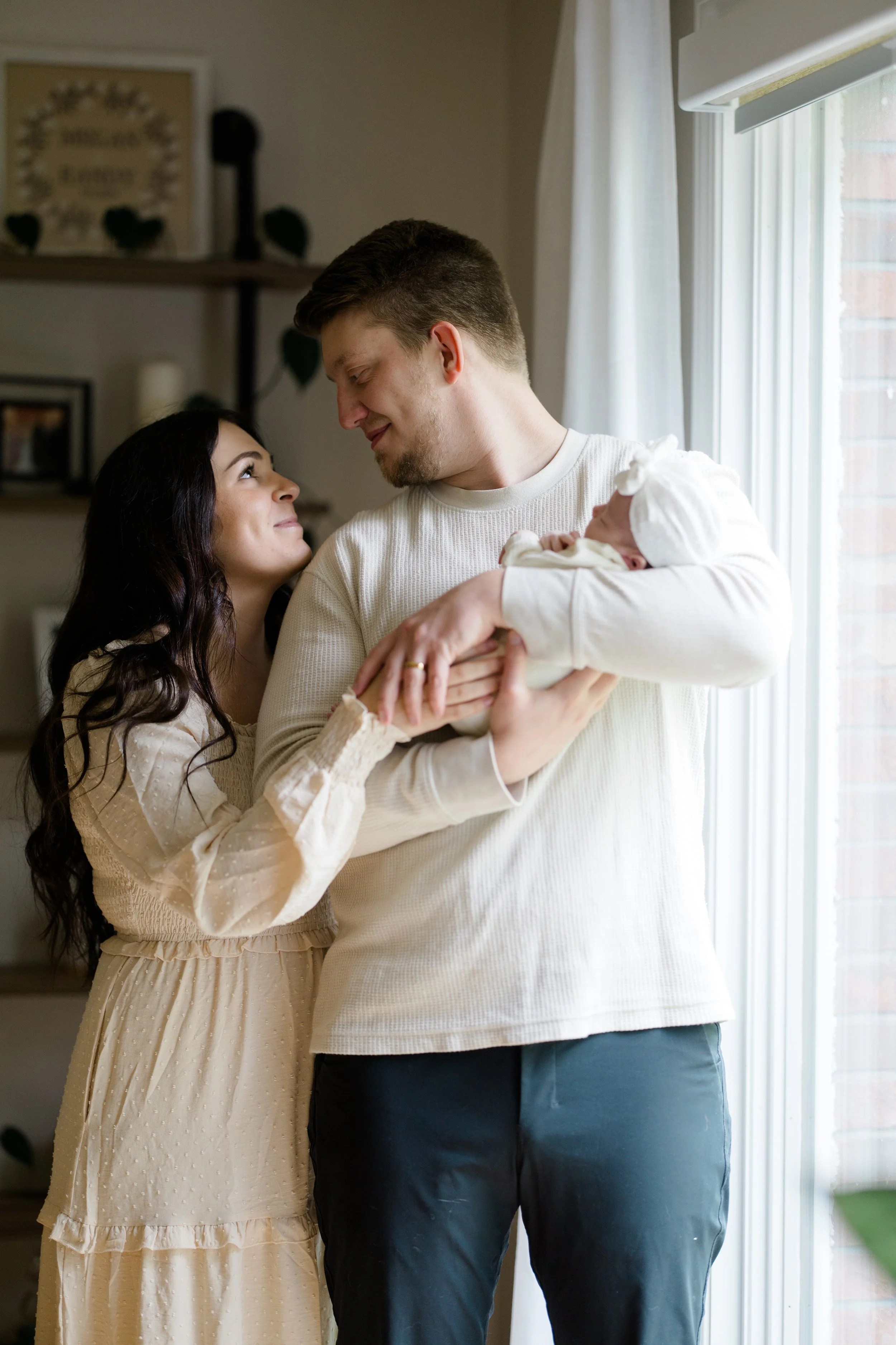 A young woman and a young man with a newborn baby, indoors near a window with white curtains, sharing a happy moment, both smiling and looking at each other.