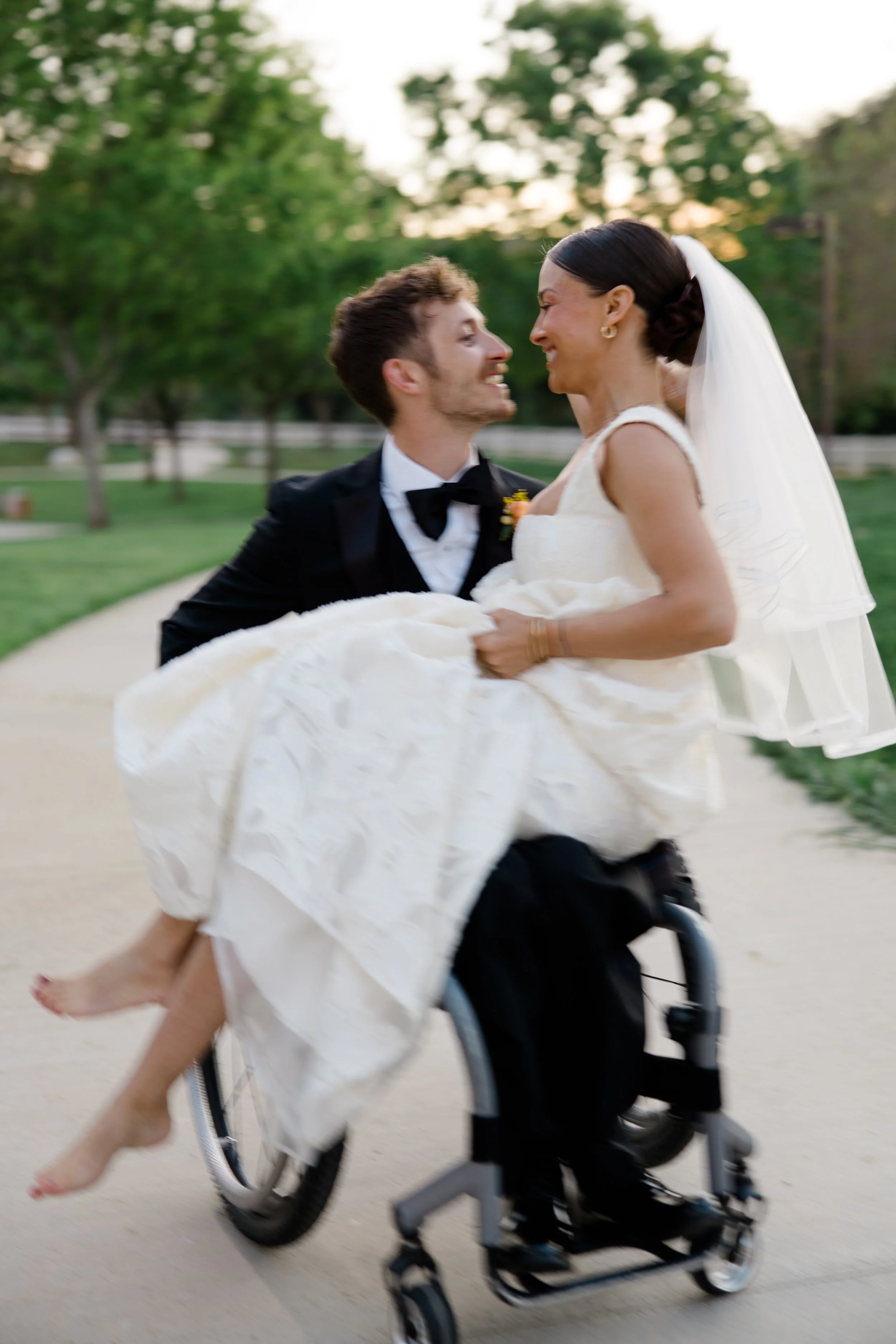 A man and woman sharing a joyful moment, with the man in formal attire and the woman in a wedding dress, as he carries her in a wheelchair outdoors during sunset.