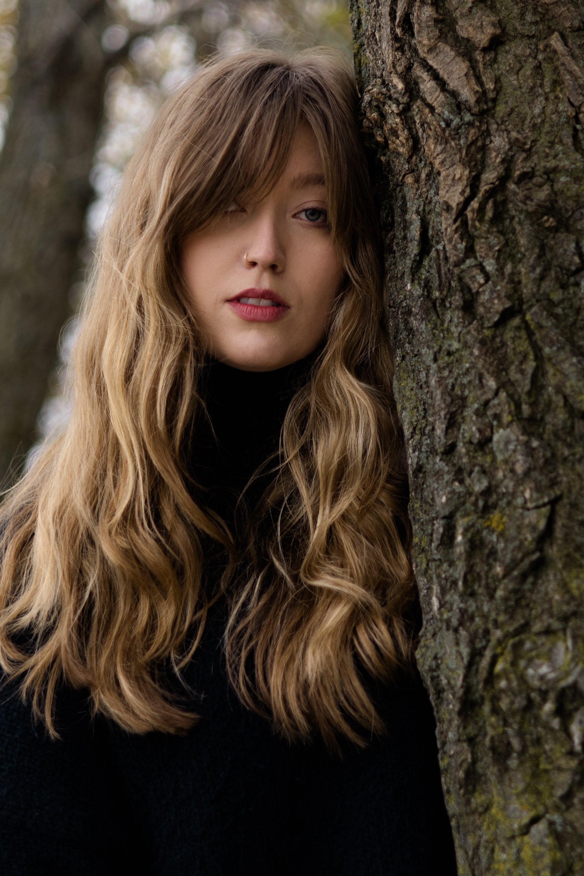 A young woman with long wavy hair and a nose piercing, partially hidden behind a tree trunk, outdoors.