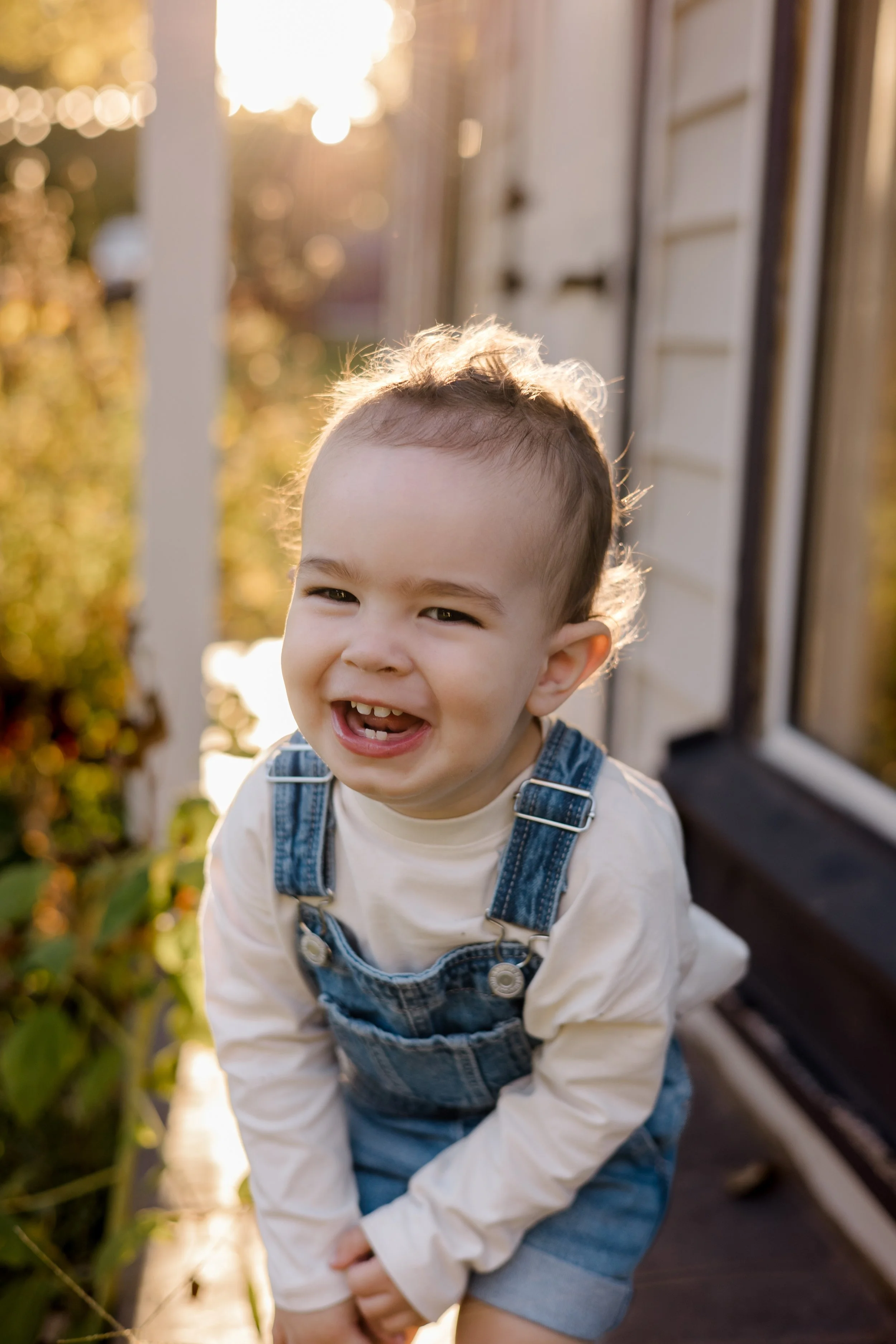 A young child with curly hair smiling and squatting outside in a garden, wearing a white shirt and denim overalls, illuminated by warm sunlight.