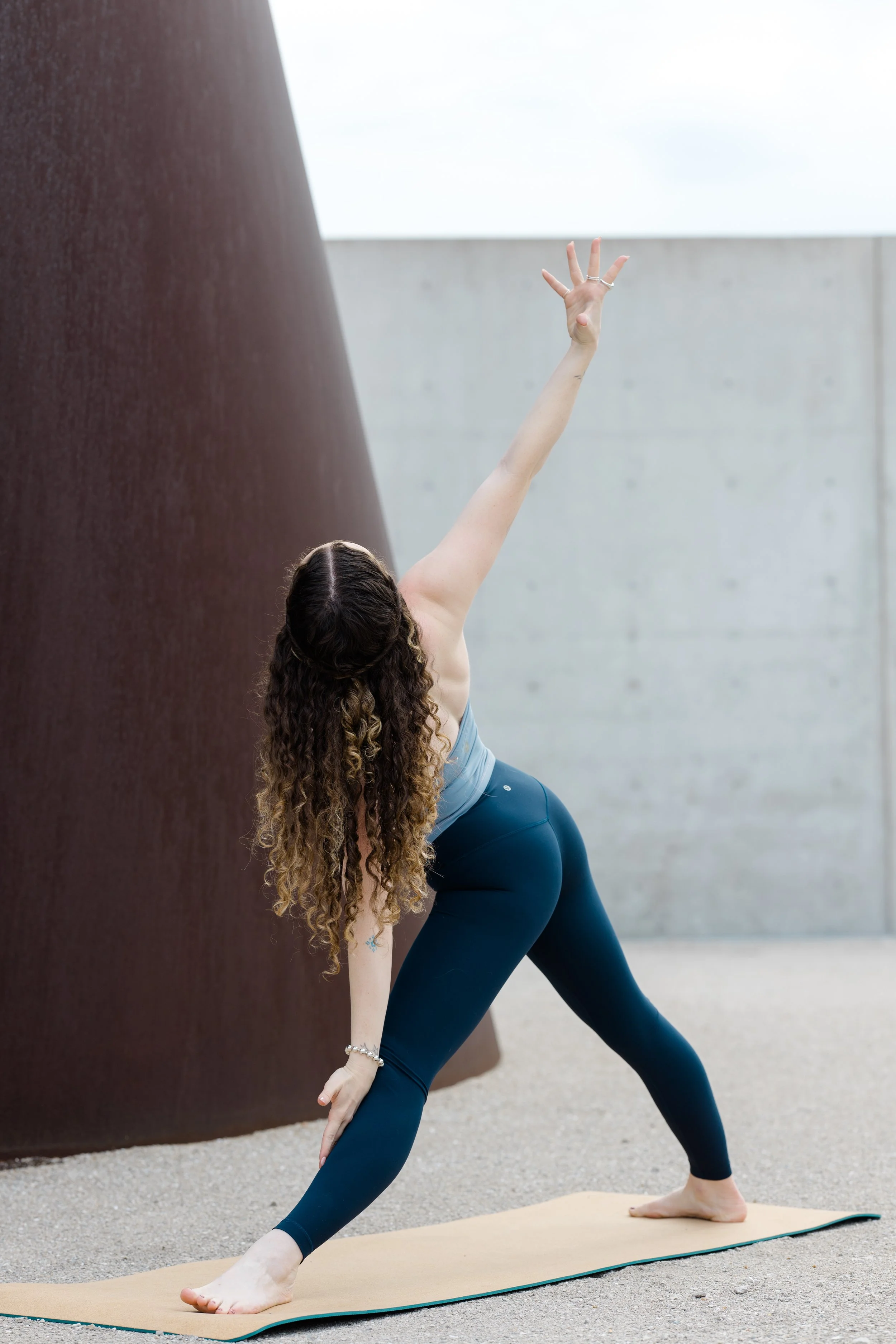 A woman with long curly hair practicing yoga outdoors on a beige mat, wearing blue leggings and a light top, stretching forward with her left hand on her shin and her right hand reaching up, against a backdrop of a large concrete wall and a cloudy sk