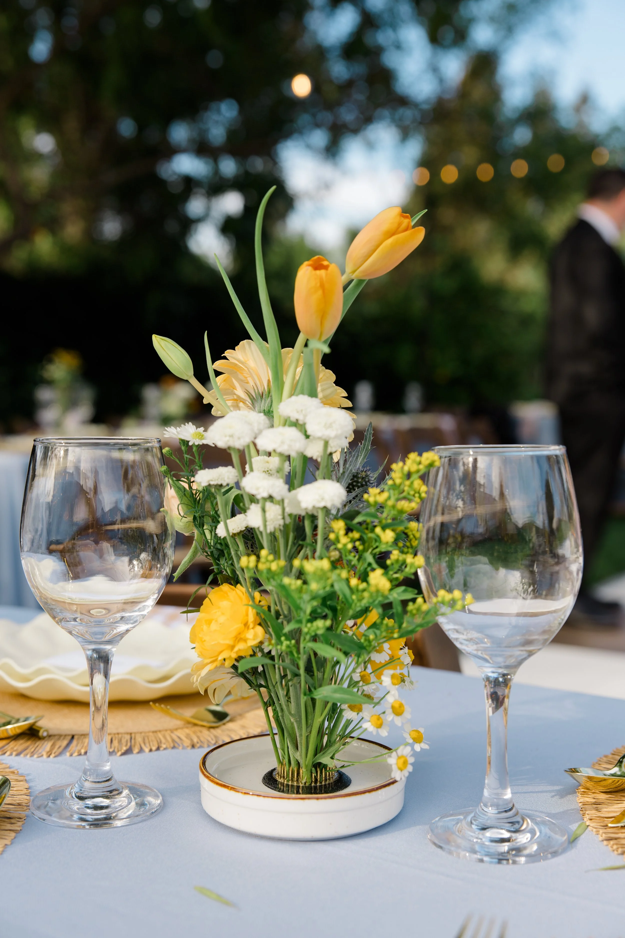 Elegant table centerpiece with yellow and white flowers, two empty wine glasses, and gold-colored placemats on a white tablecloth outdoors with blurred greenery background.