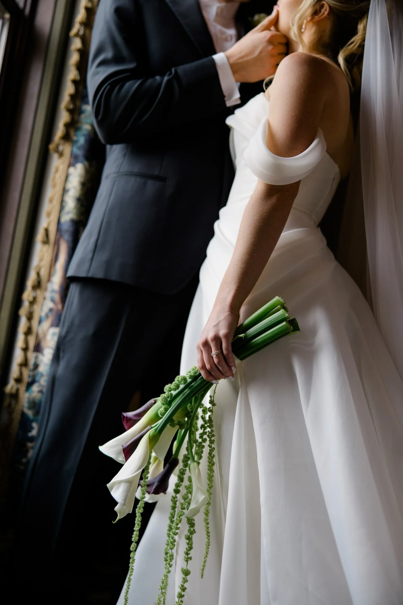 A bride in a white wedding dress holding a bouquet of calla lilies with green accents at her side, standing next to a person in a dark suit, in an ornate room.