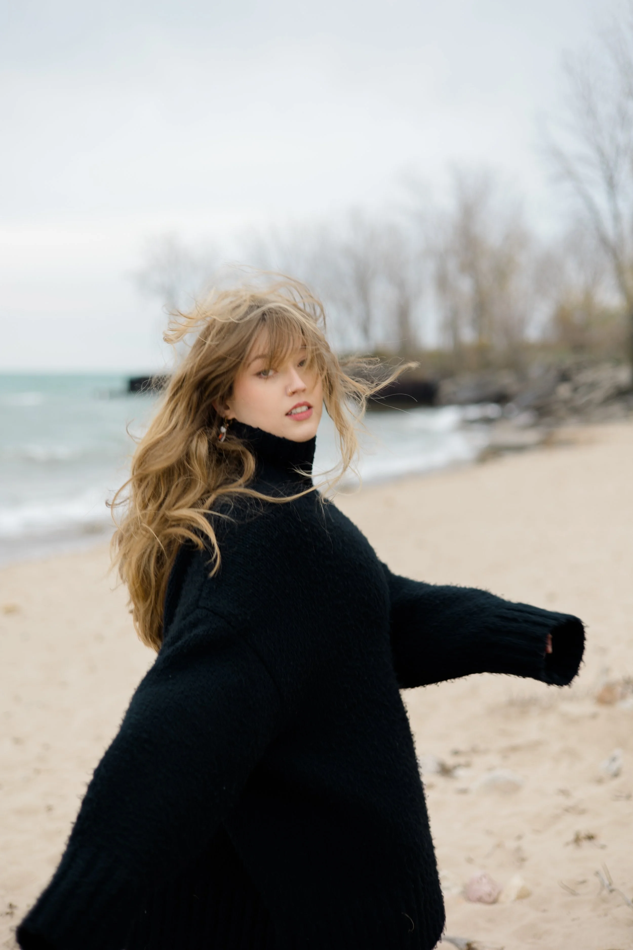 A young woman with long, wavy blonde hair stands on a sandy beach with the ocean and leafless trees in the background. She is wearing a black coat and looks at the camera with a slight smile.