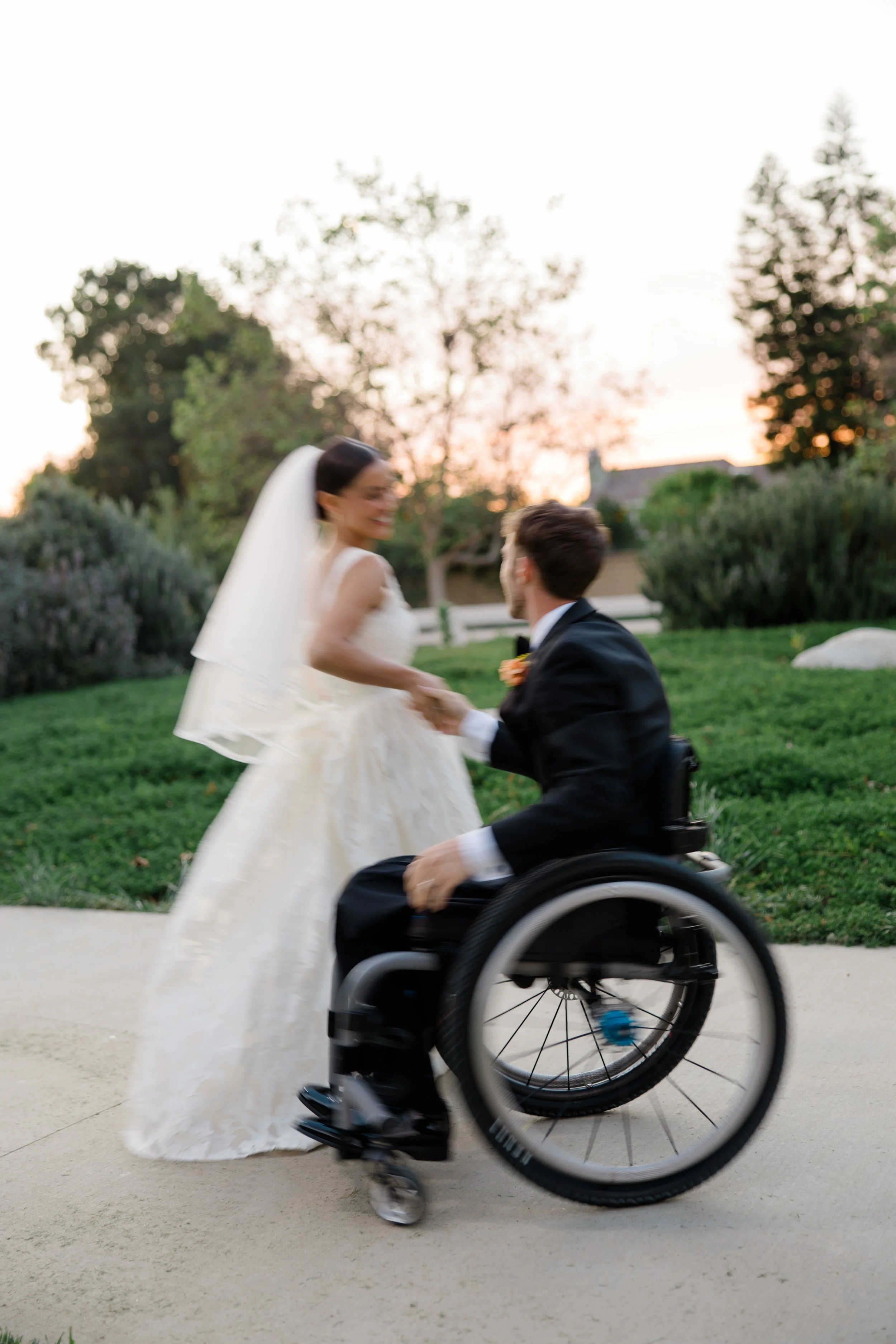 A man in a wheelchair holding hands with a woman in a wedding dress outdoors at sunset, with trees in the background.