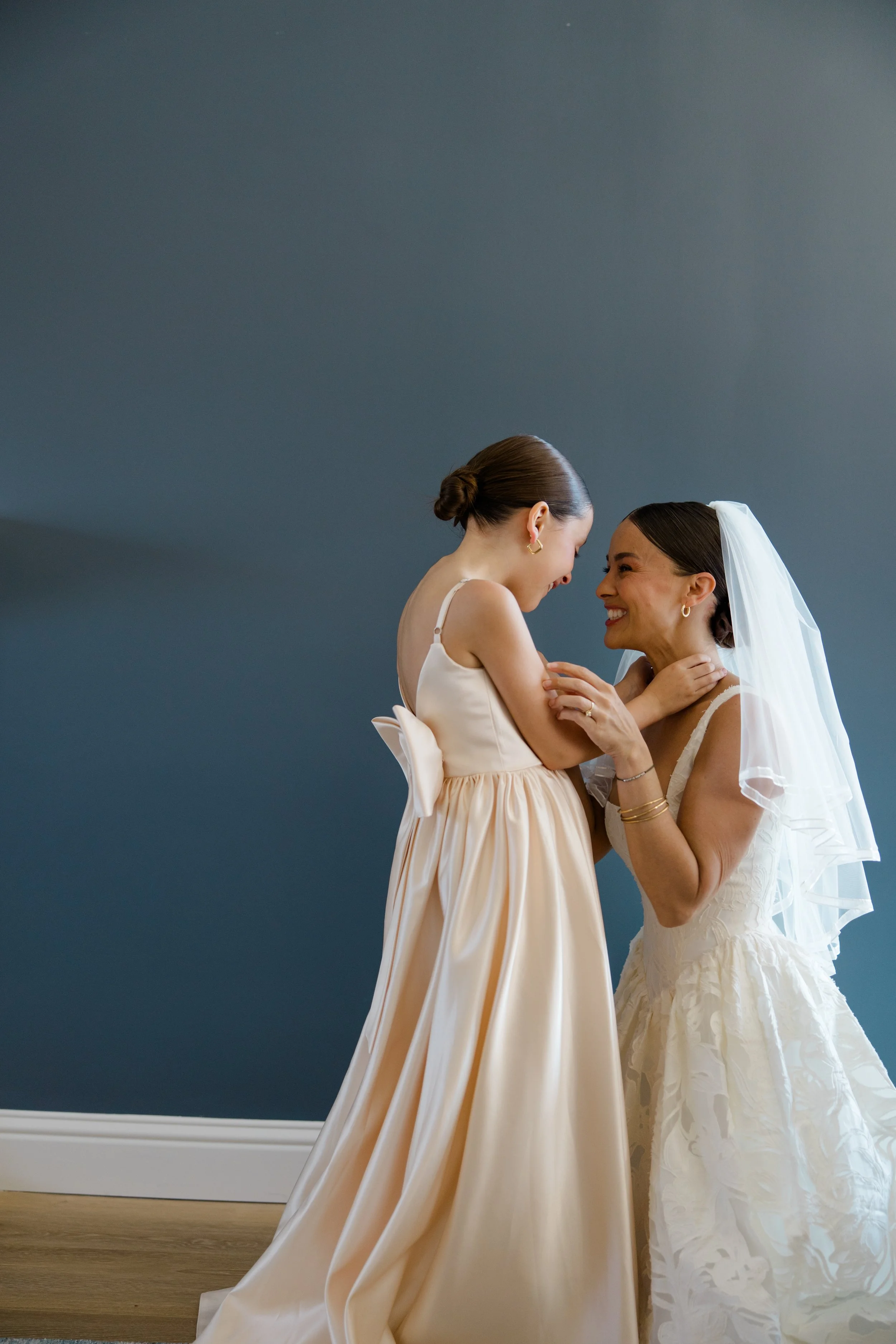 Two women on their wedding day smiling and touching each other lovingly. One bride is wearing a white wedding dress with a lace bodice and veil, and the other woman in a cream-colored dress with a large bow on the back. They are indoors against a dar