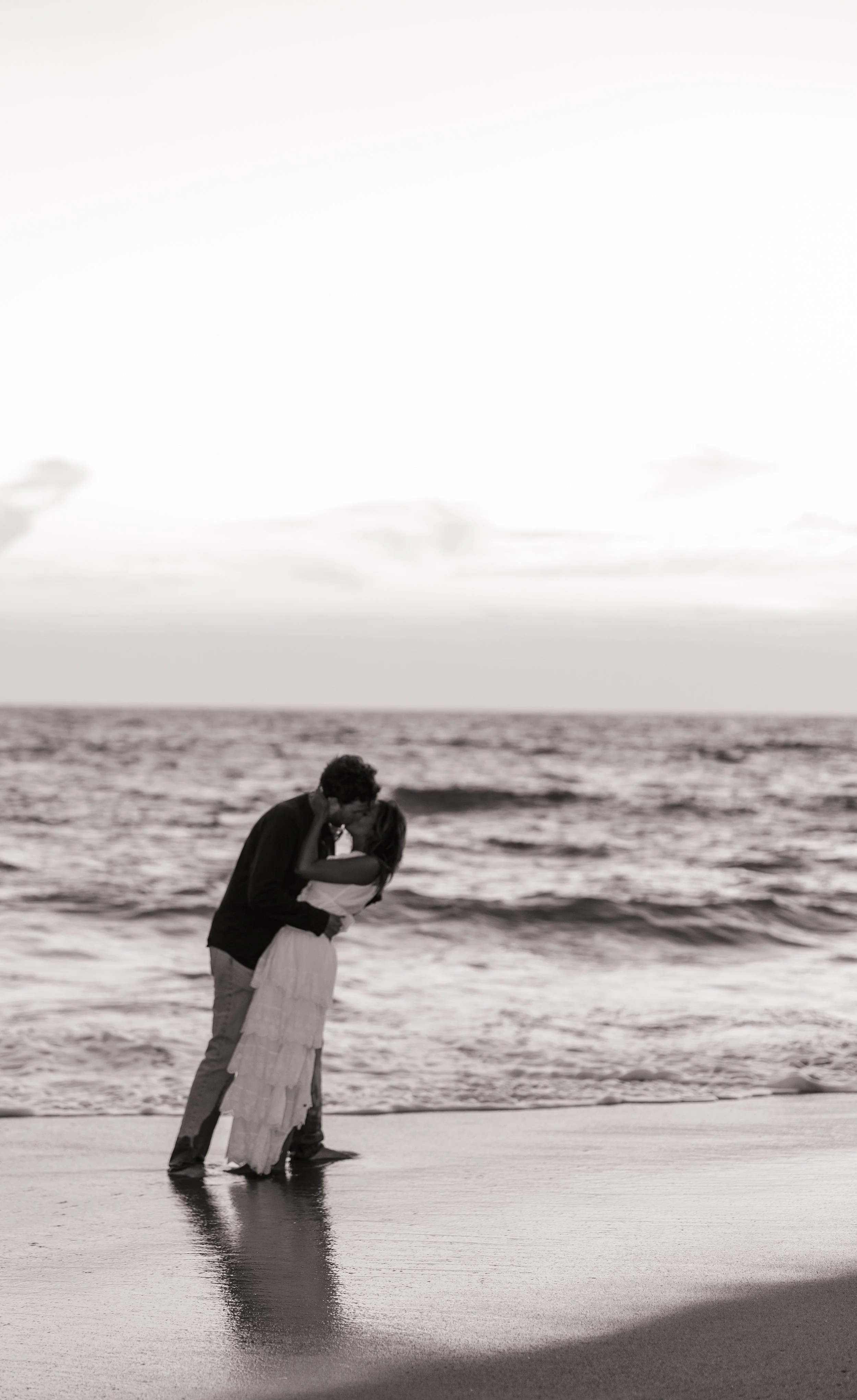 A couple in love sharing a kiss on the beach, standing in the shallow water with the ocean and cloudy sky in the background.