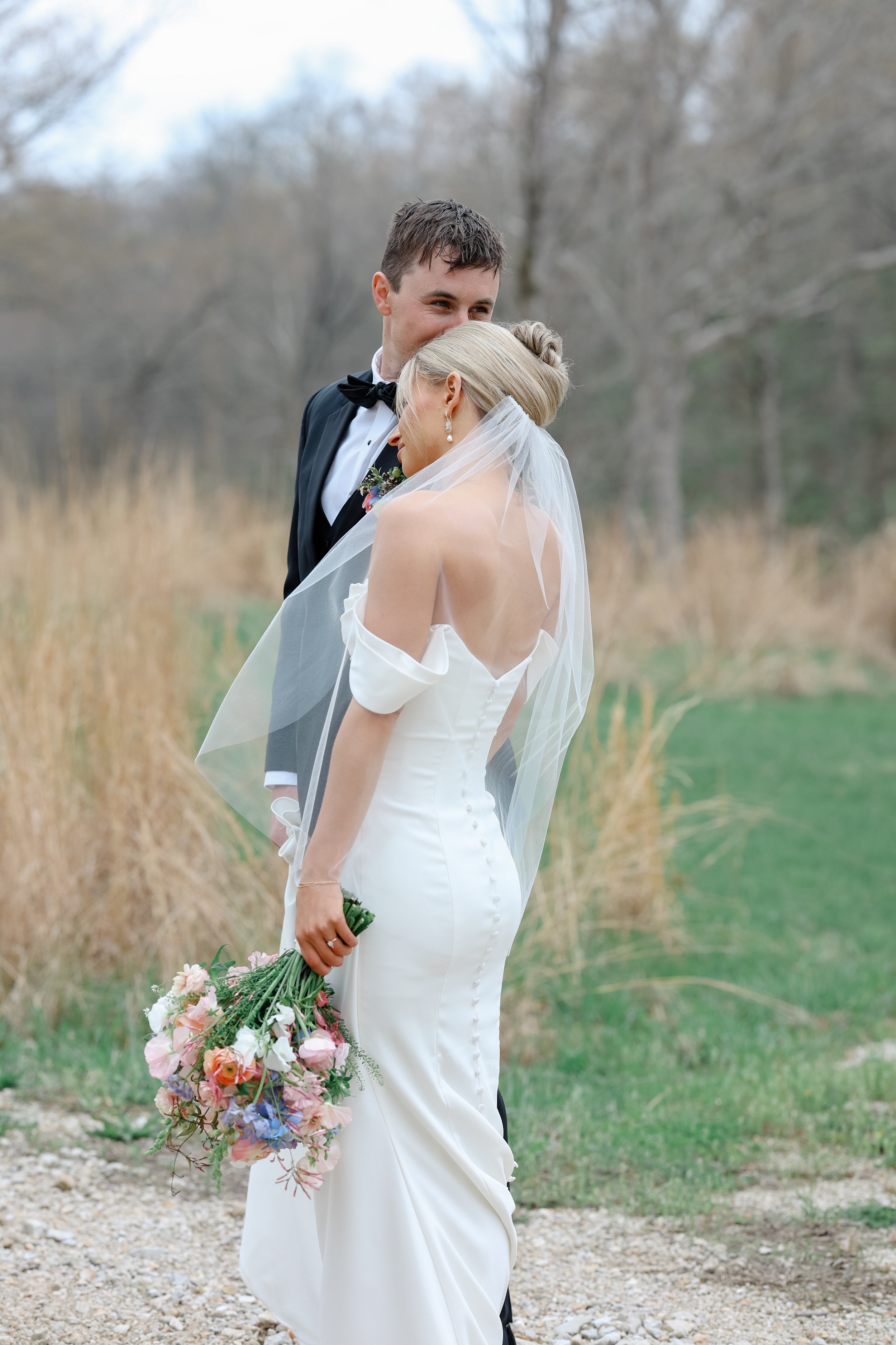 A bride and groom standing close together in an outdoor setting with bare trees and grass. The bride is holding a bouquet of colorful flowers and has a veil, and is wearing a white off-shoulder wedding dress. The groom is in a black tuxedo with a bow