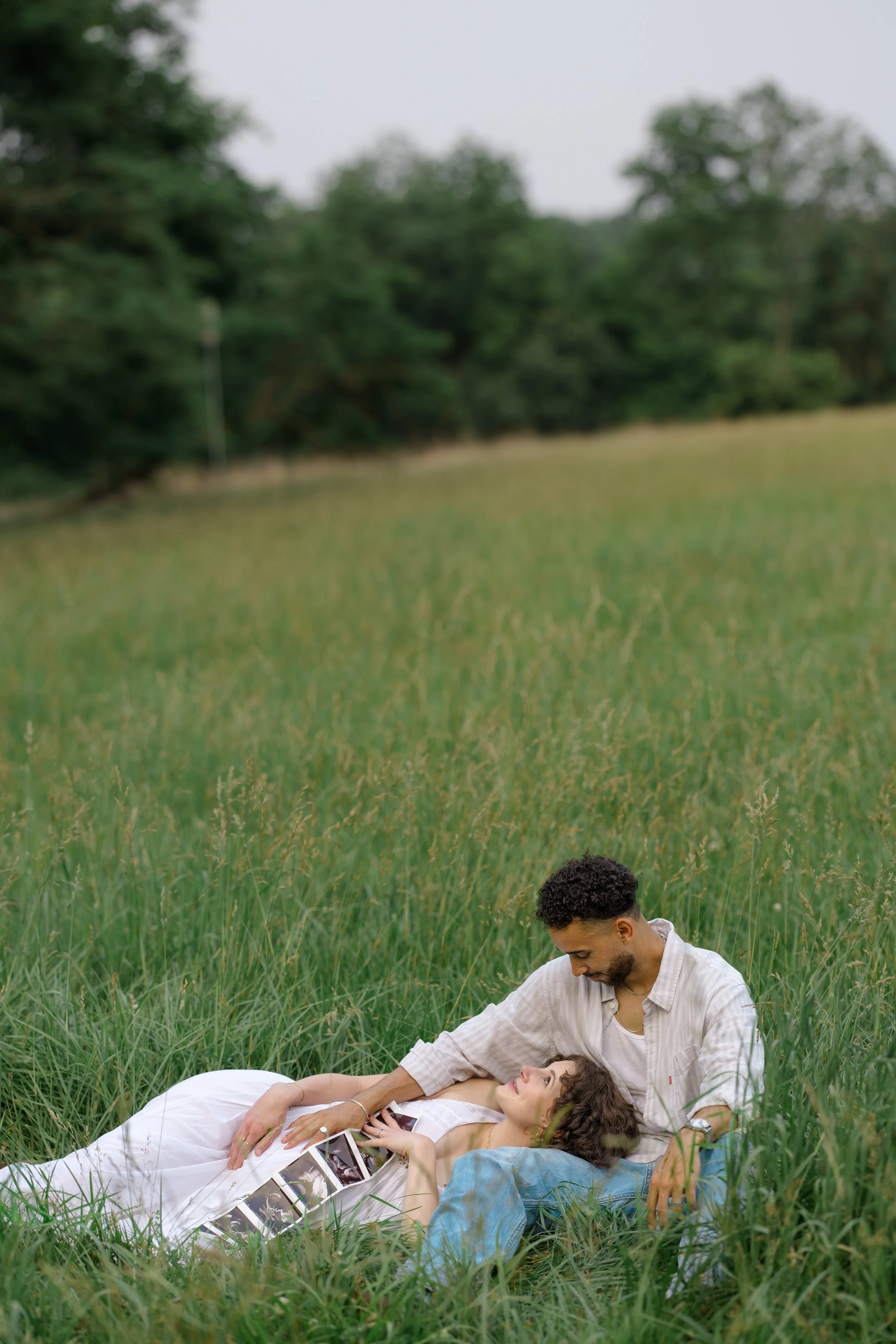 A man and a woman lying on the grass in a field with tall grass and trees in the background. The woman is lying down holding photographs, while the man is sitting beside her, leaning over her, and looking at her.
