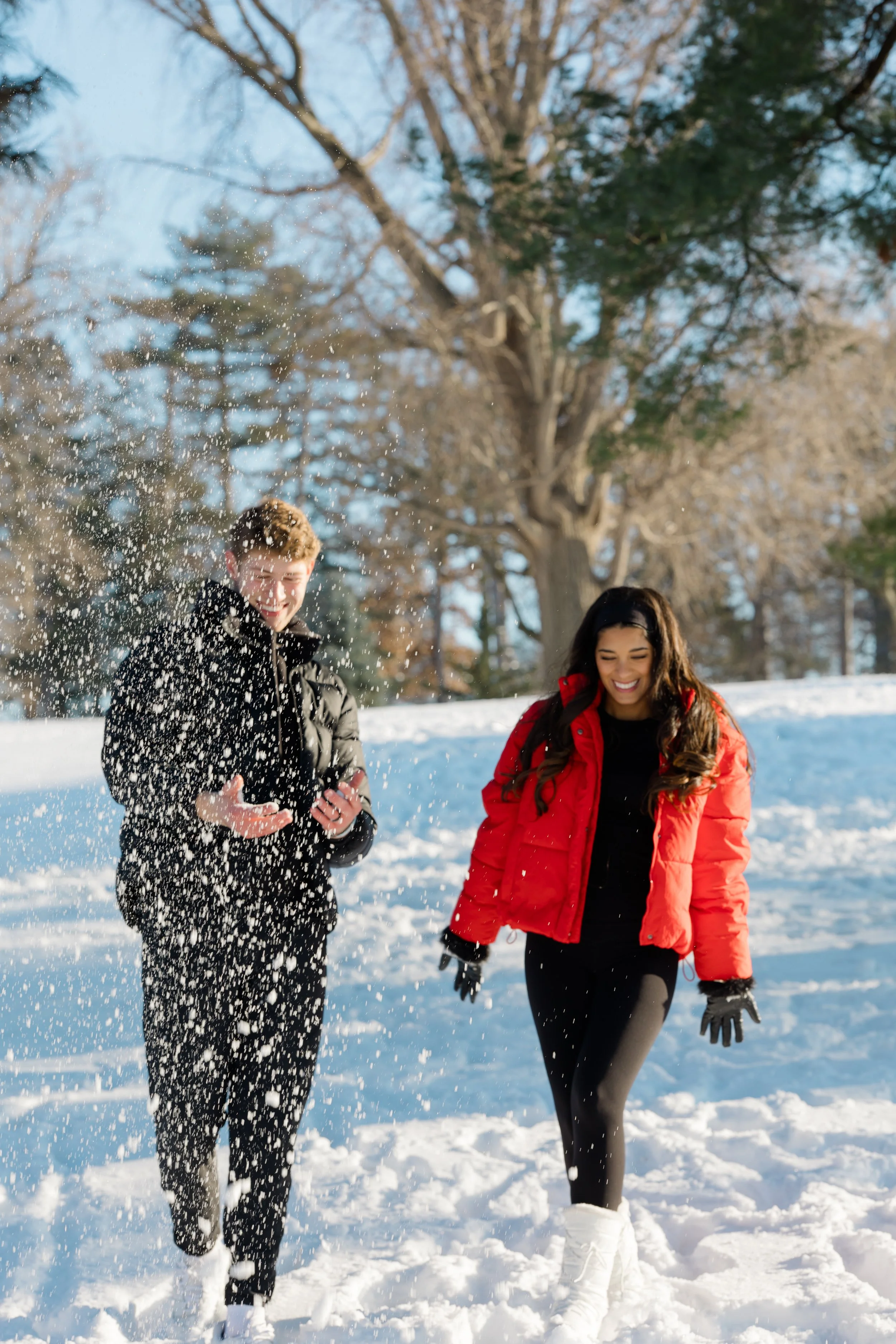 Two young adults, one male and one female, joyfully playing in the snow during winter, with leafless trees in the background.
