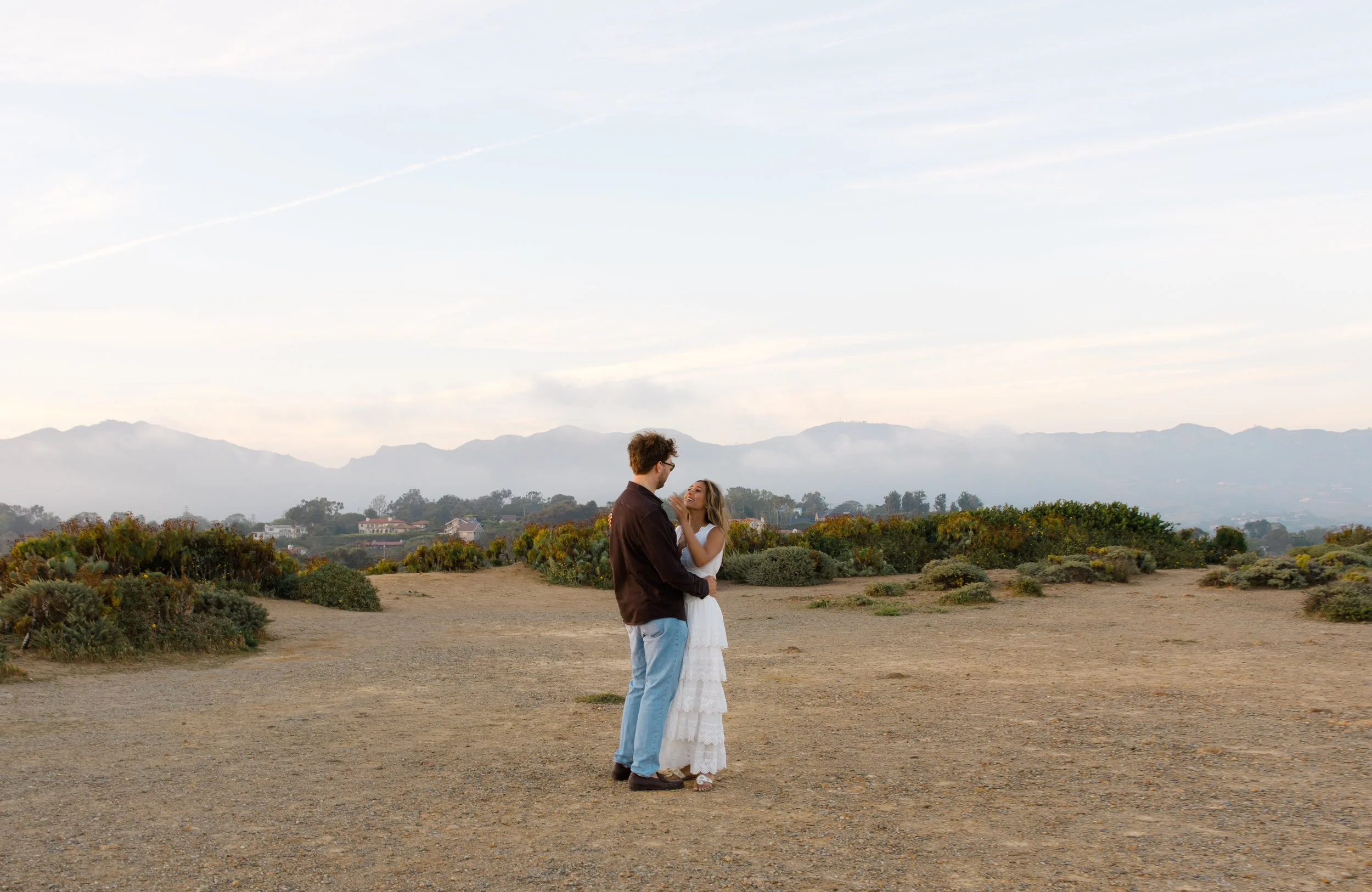 A couple standing close together outdoors on a dirt path, holding each other, with mountains in the background under a cloudy sky.