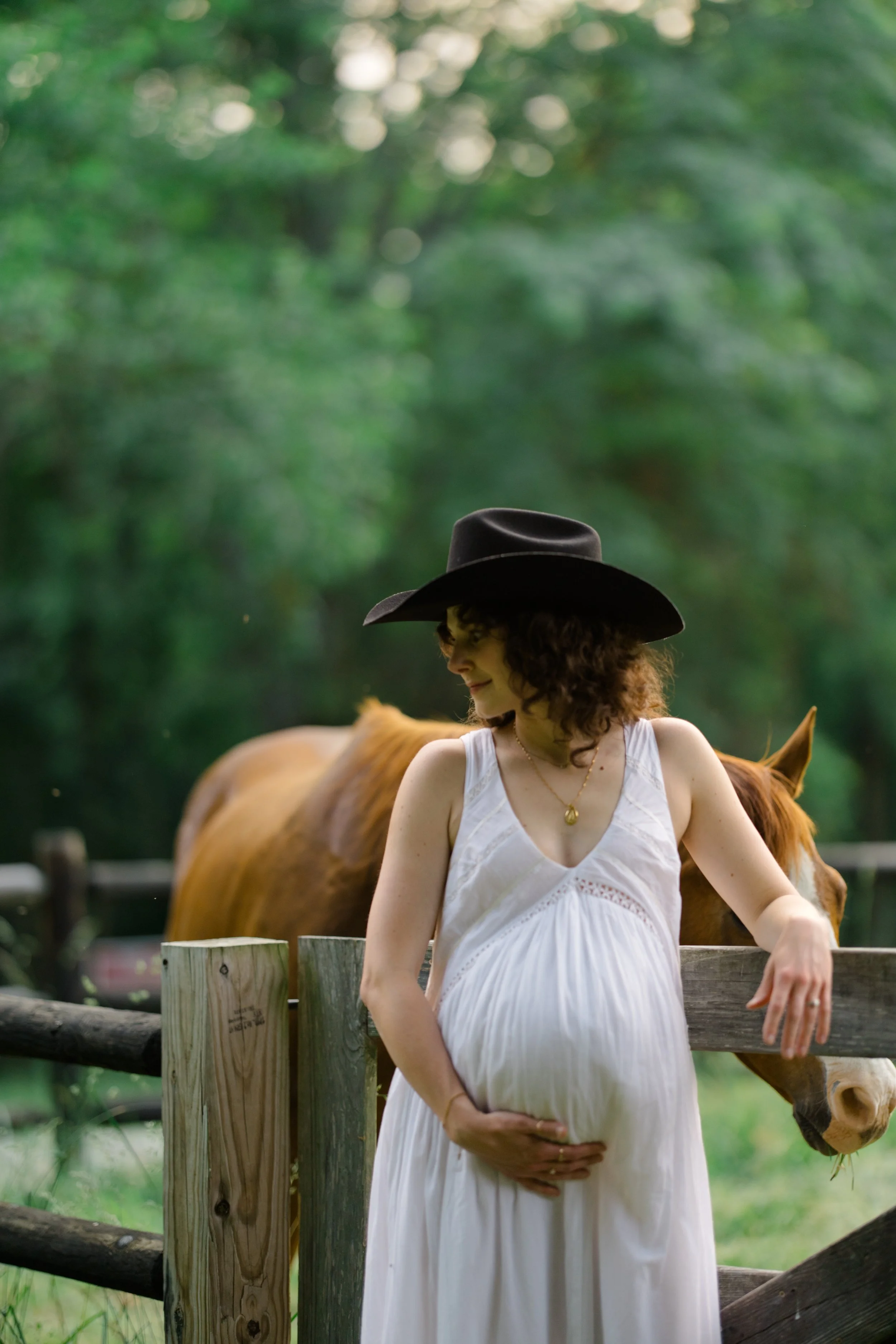 A woman in a white dress and a black wide-brimmed hat stands outdoors by a wooden fence, with a brown horse in the background on a lush green day.