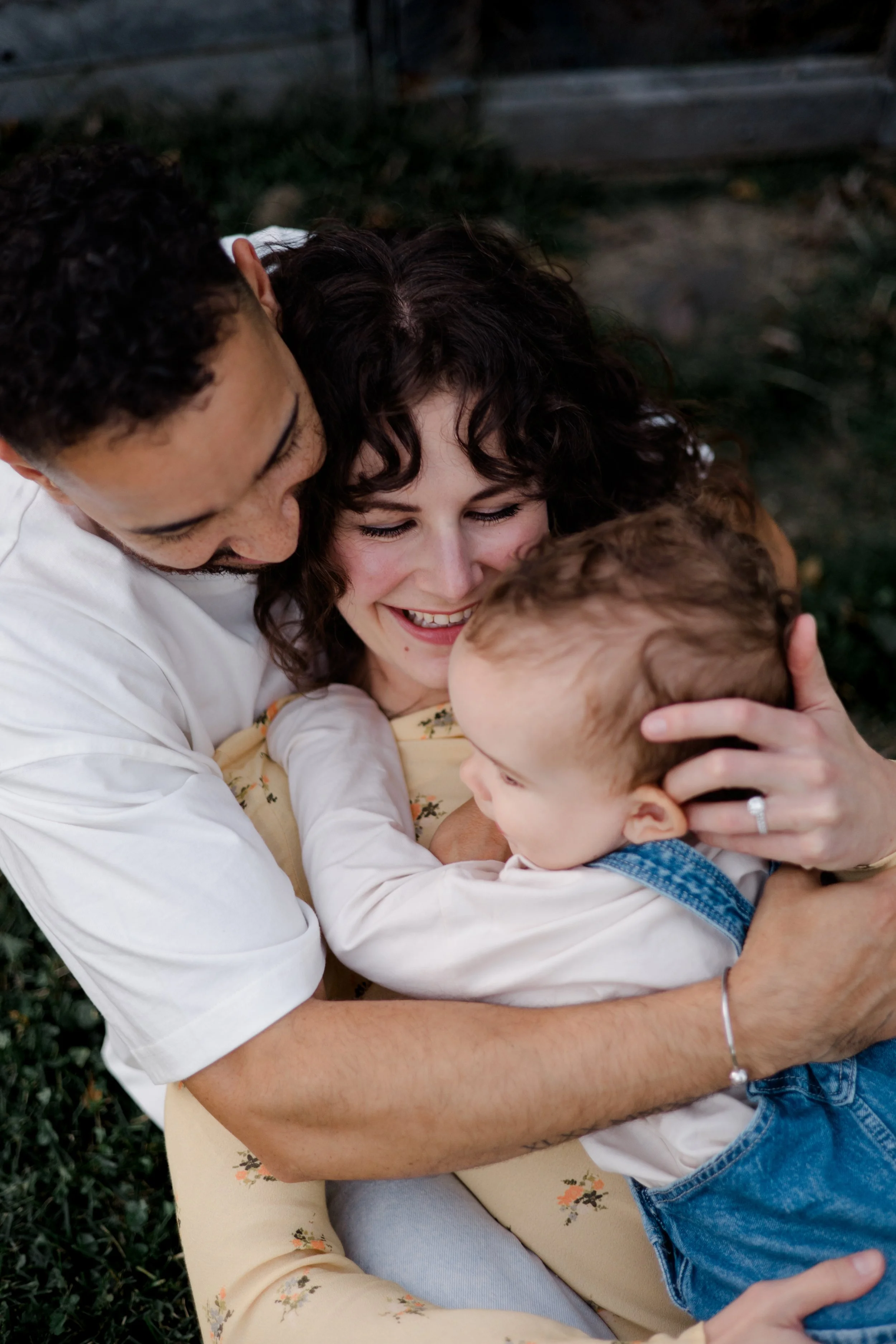 A family hug, with a woman with curly dark hair, a man with short curly hair, and a young boy with light brown hair, all smiling and embracing each other outdoors.