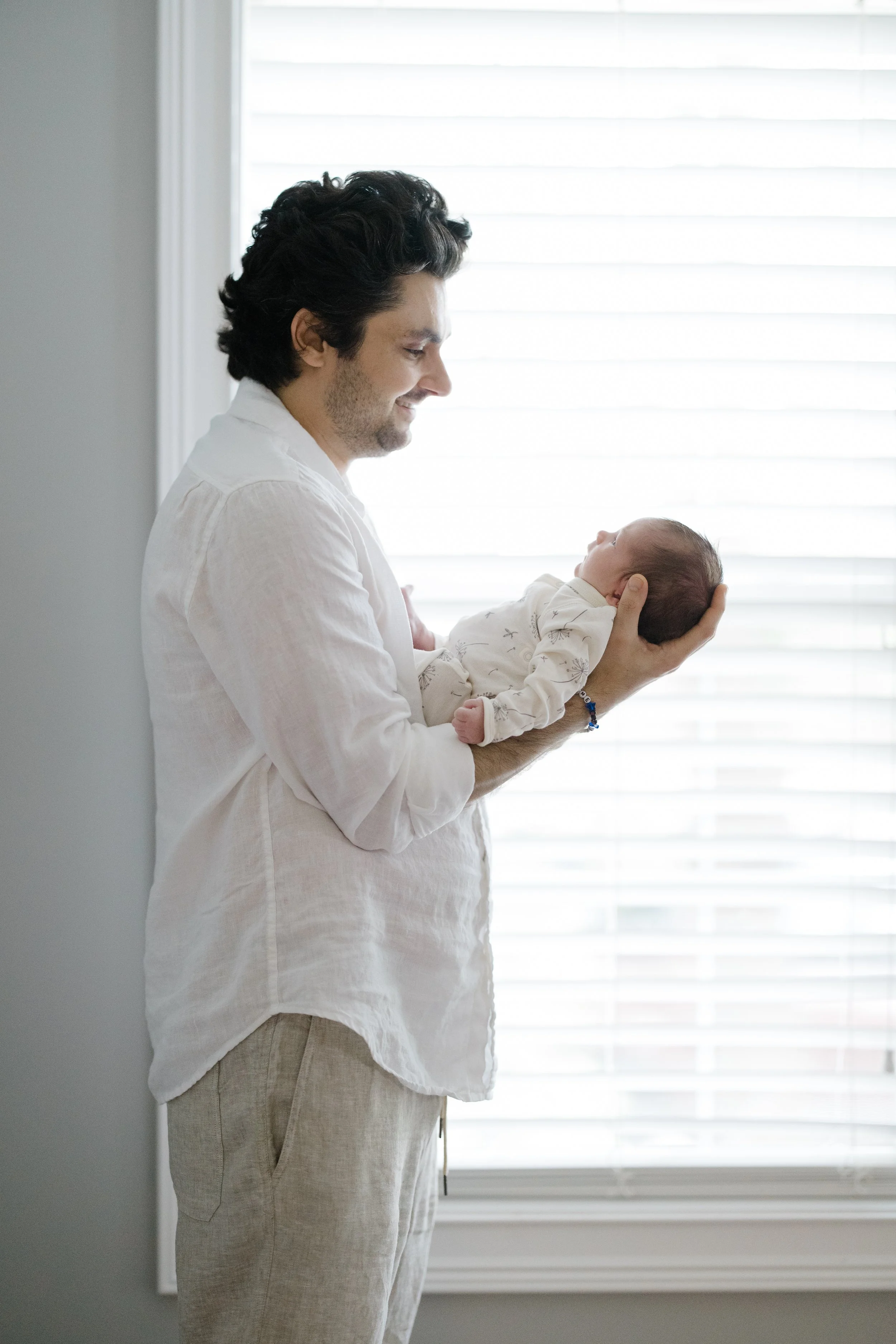 A man with dark curly hair and a beard, wearing a white shirt and beige pants, holds a newborn baby in his hands while standing near a window with white blinds, smiling softly at the baby.