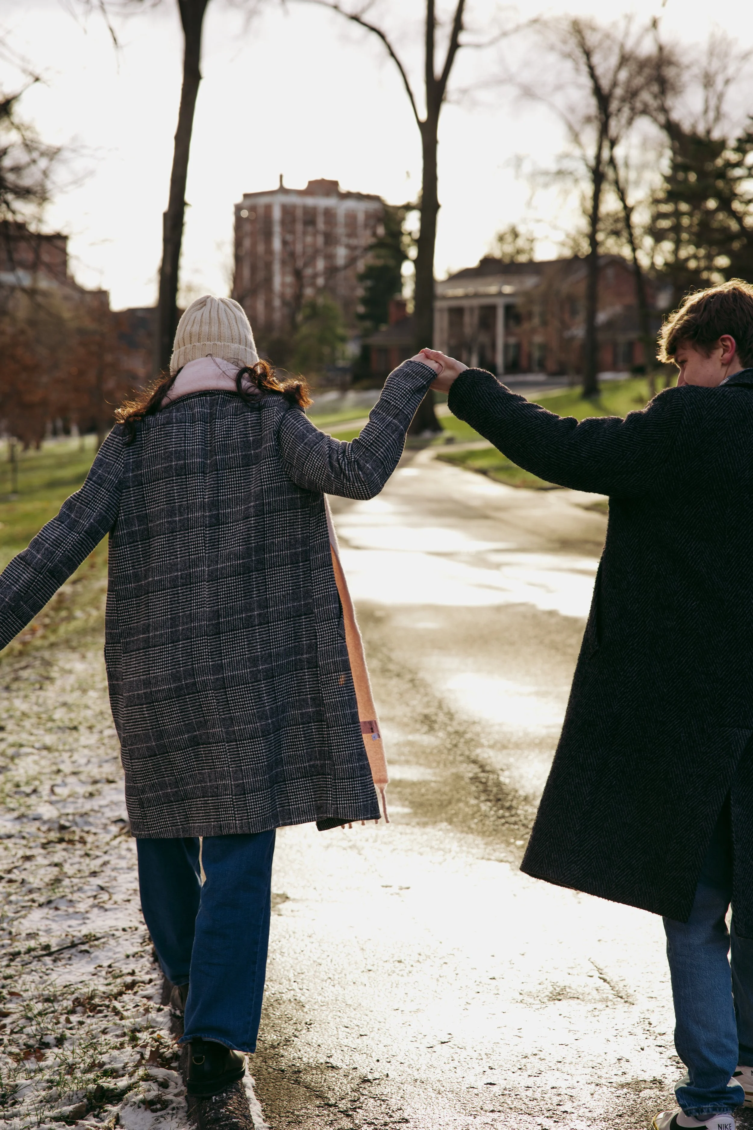 Two people holding hands while walking outdoors on a sidewalk in a park during daytime, with trees and buildings in the background.