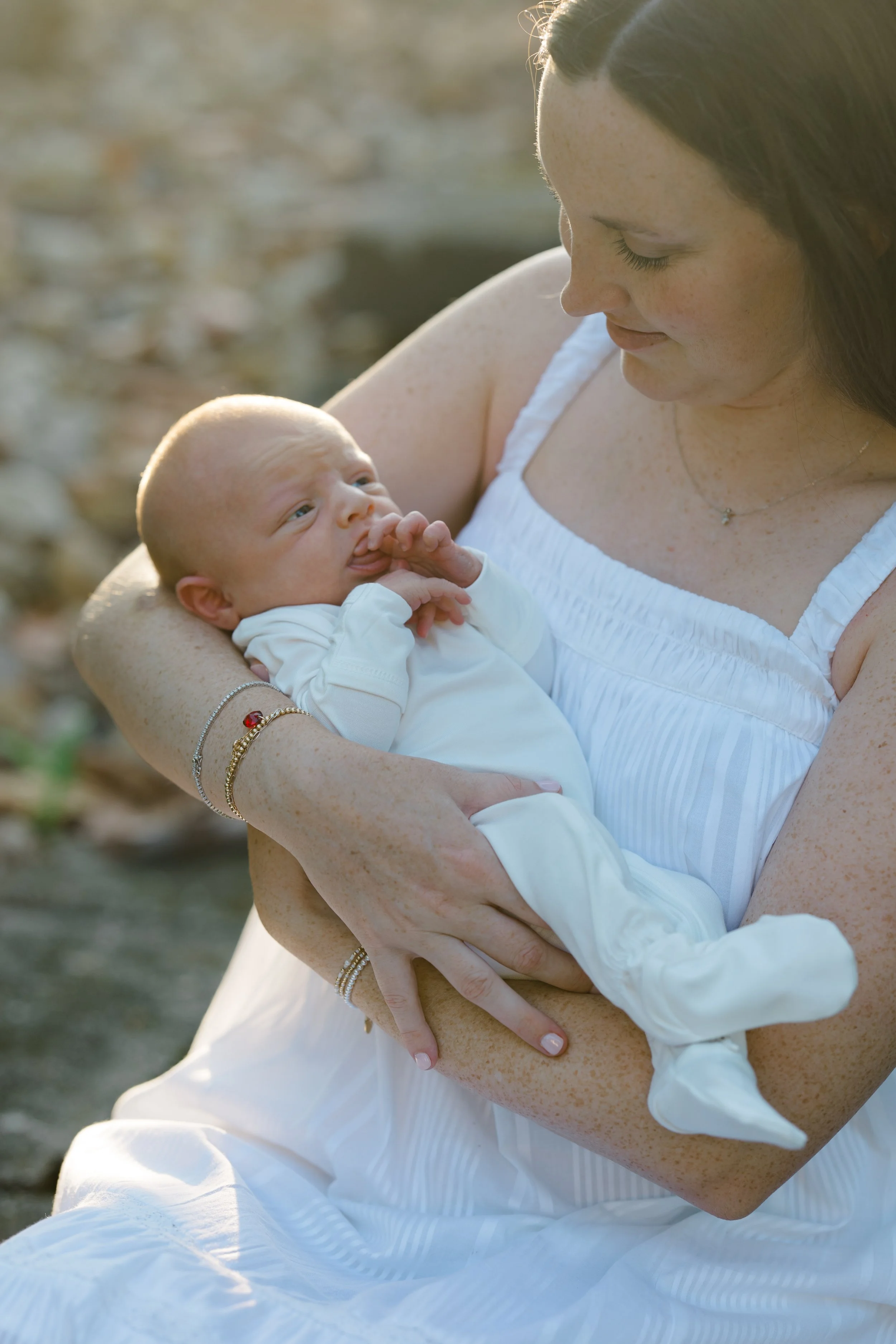 A woman with long brown hair, freckles, and wearing a white dress holds a newborn baby dressed in white, gazing at the baby with a gentle expression.