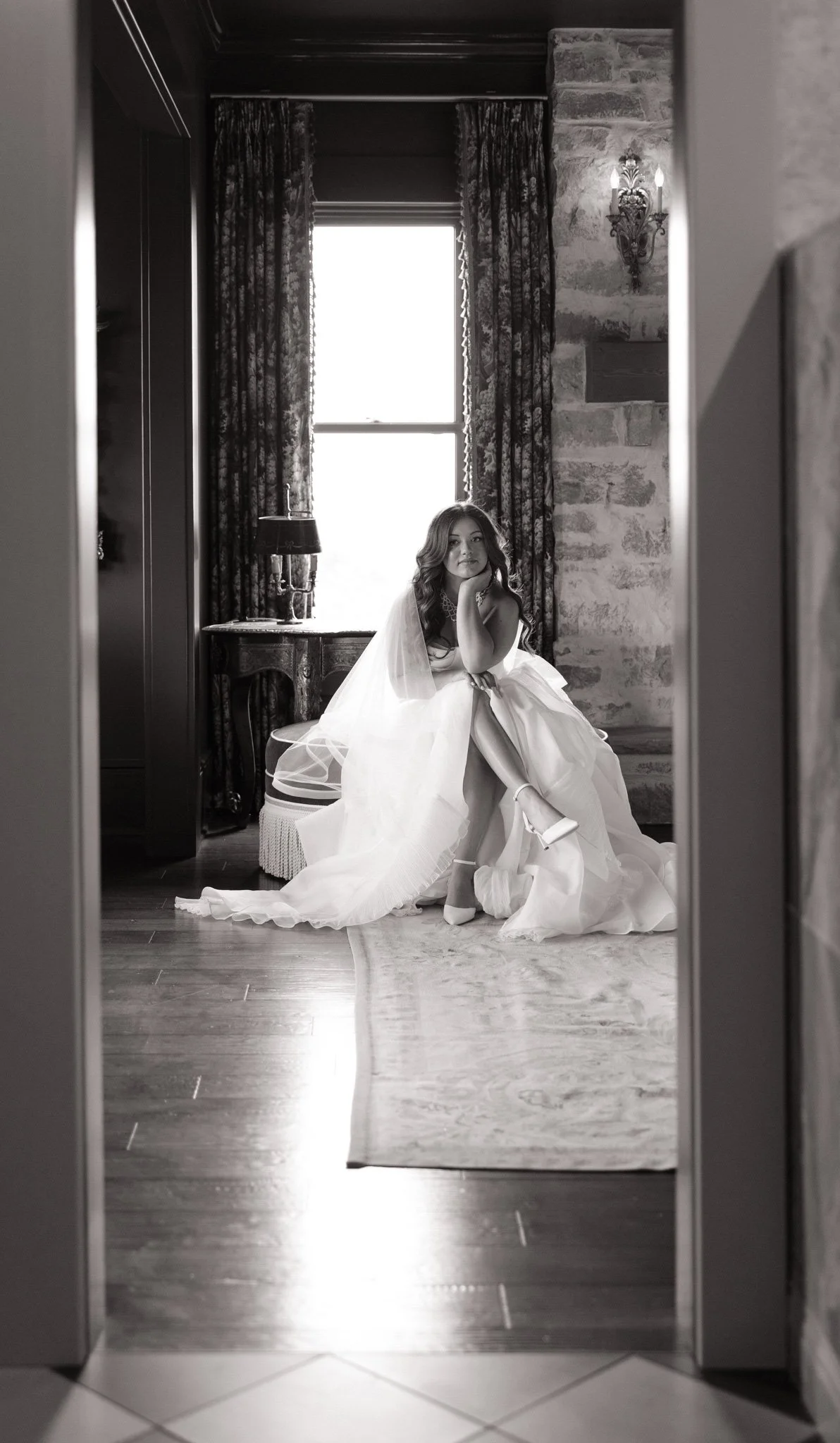 A woman in a wedding gown sitting on a bench, looking at the camera, in a room with large window, curtains, stone walls, and ornate wall sconce lighting, viewed through a doorway.
