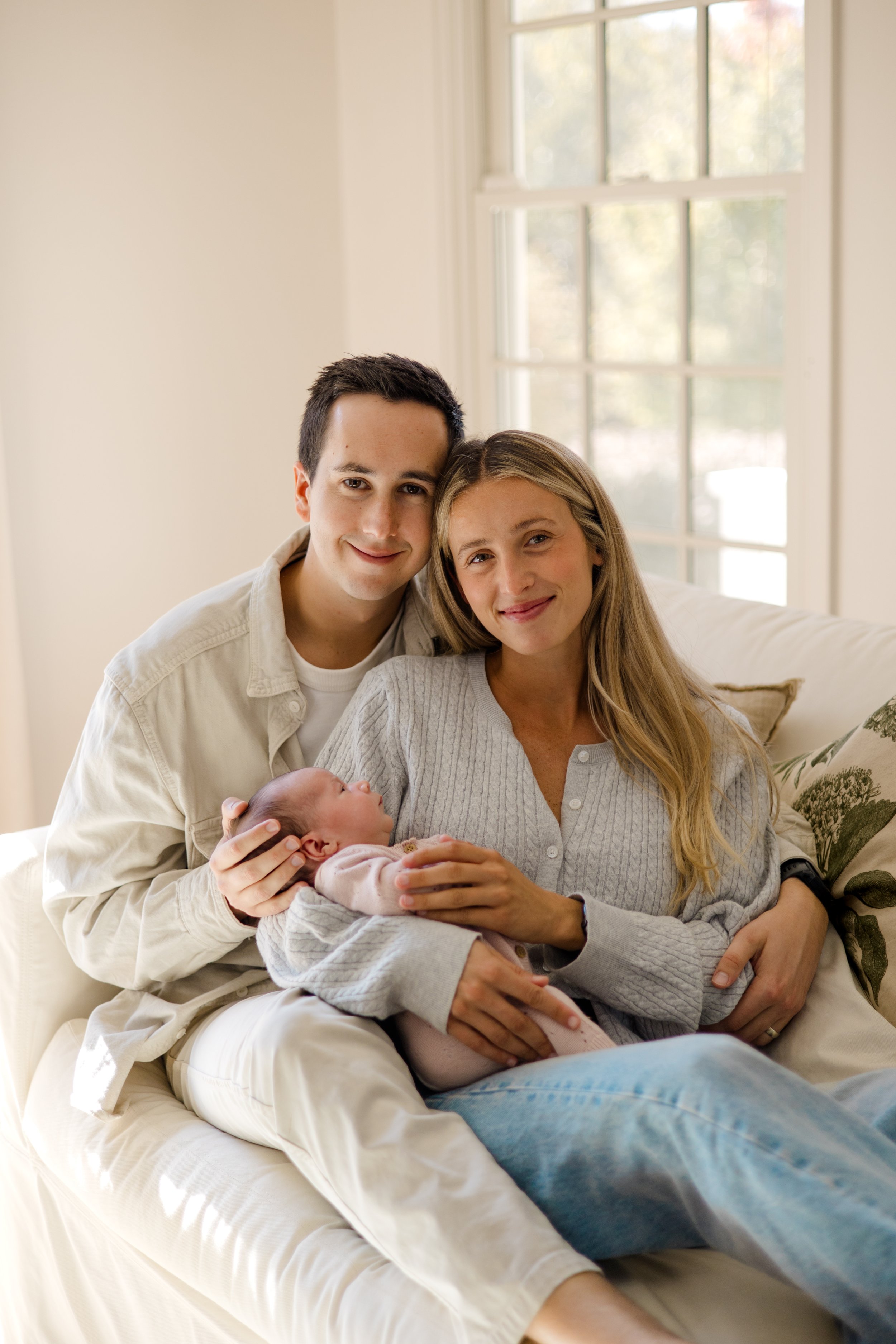 A multi-generational family sitting together on a cream-colored couch in a bright room with large windows, holding a newborn baby, all smiling and looking at the camera.