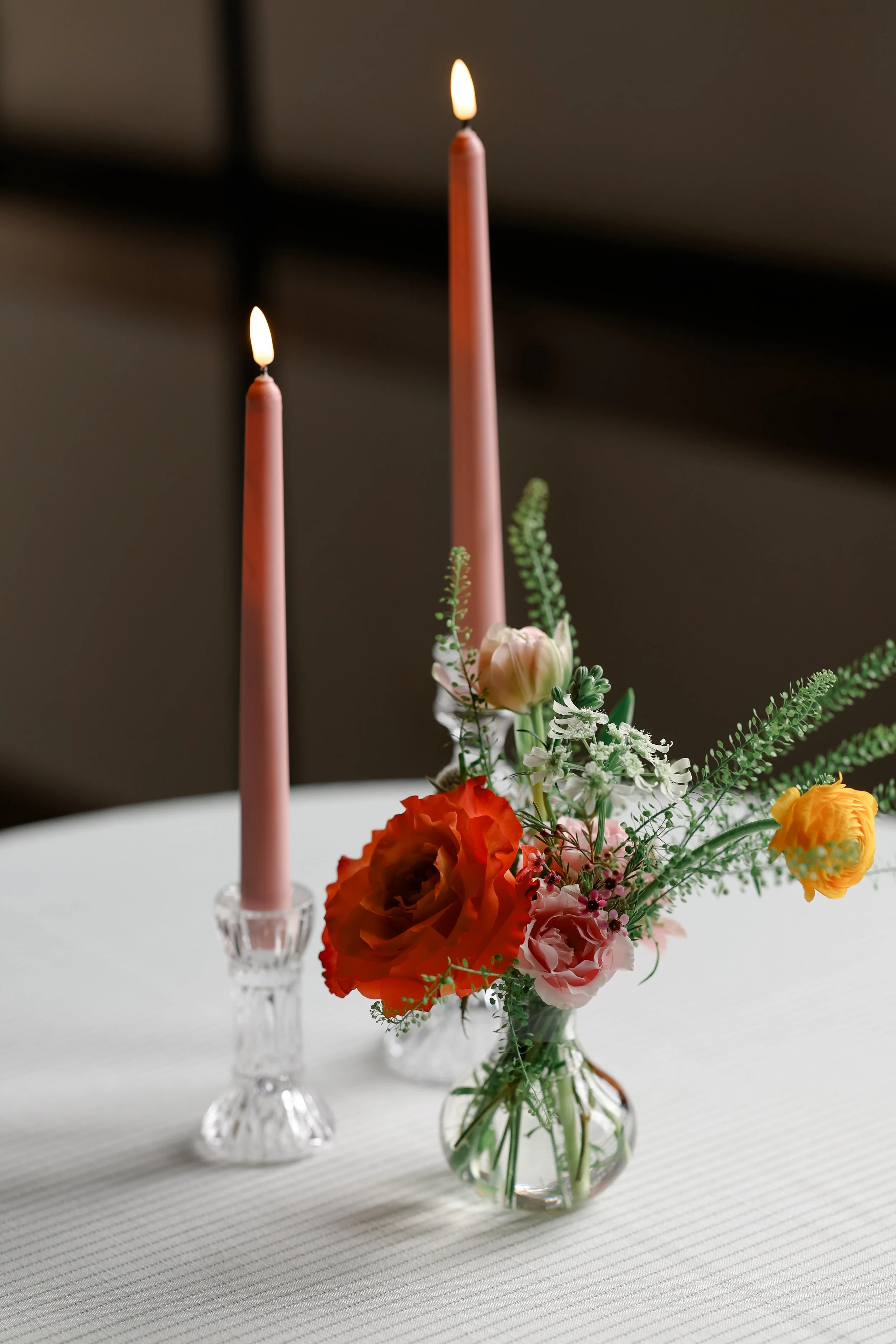 Vase with mixed flowers, including orange, pink, and yellow blooms, on a white table with two pink taper candles in clear glass holders.