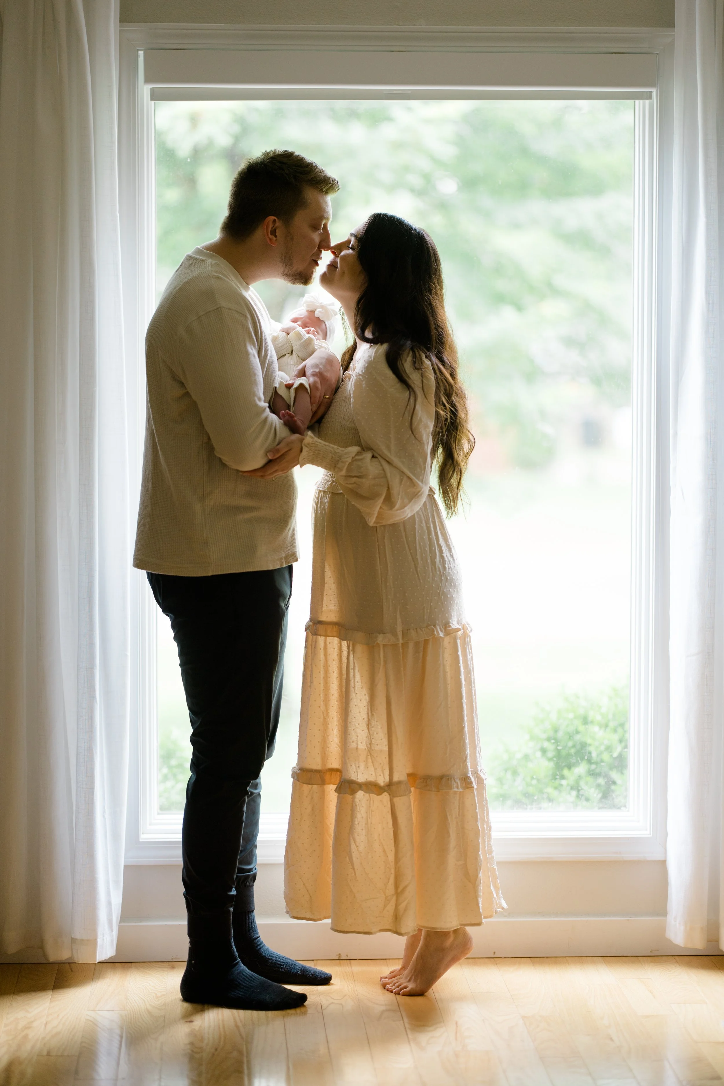 A man and woman are standing near a large window, about to kiss. The man is holding a newborn baby wrapped in a blanket. The woman is wearing a light-colored, long dress, and the man is dressed casually in a light sweater and dark pants. The scene is