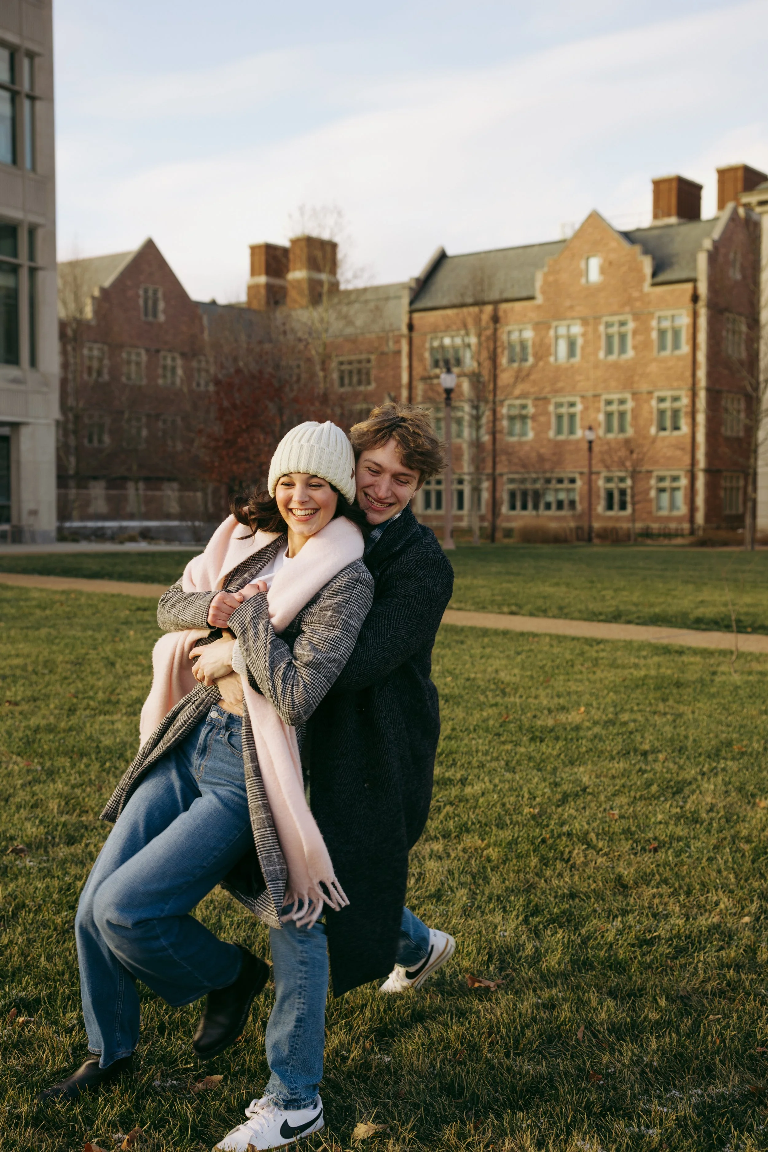 A young couple is outdoors on a grassy area, smiling and embracing each other. The woman wears a white knit hat and a pink coat, while the man wears a dark coat and both are dressed casually. In the background, there is a large brick building with mu