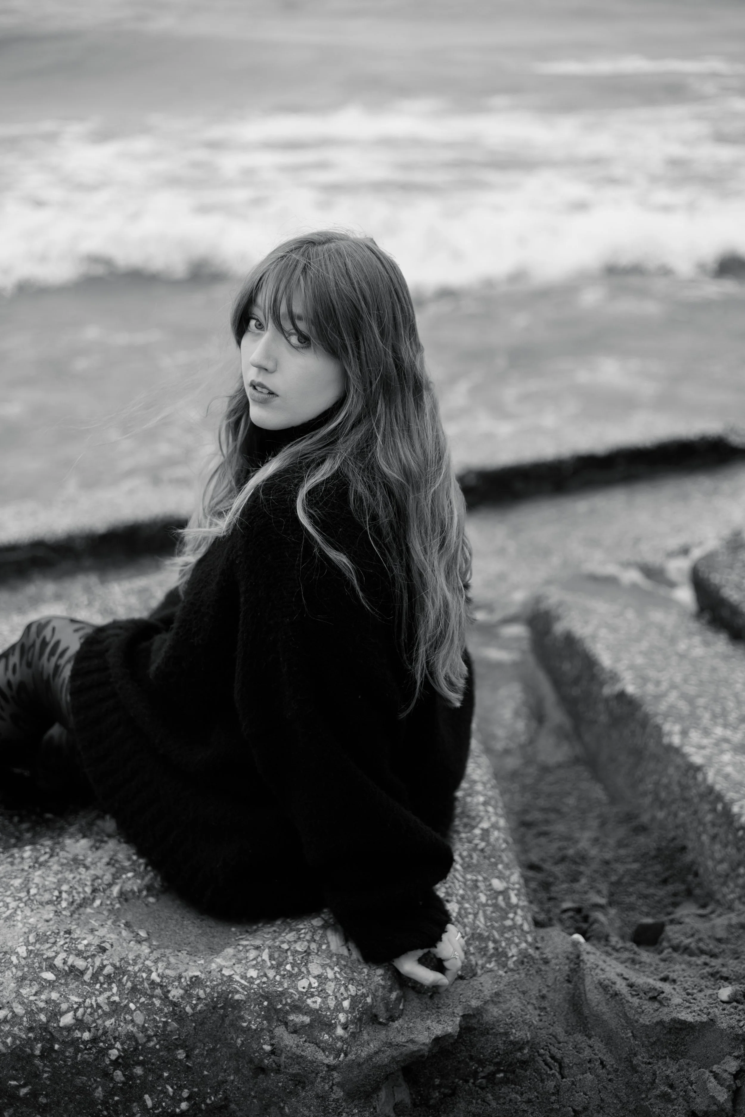 A woman with long hair sitting on a rocky beach, looking at the camera, with waves in the background.