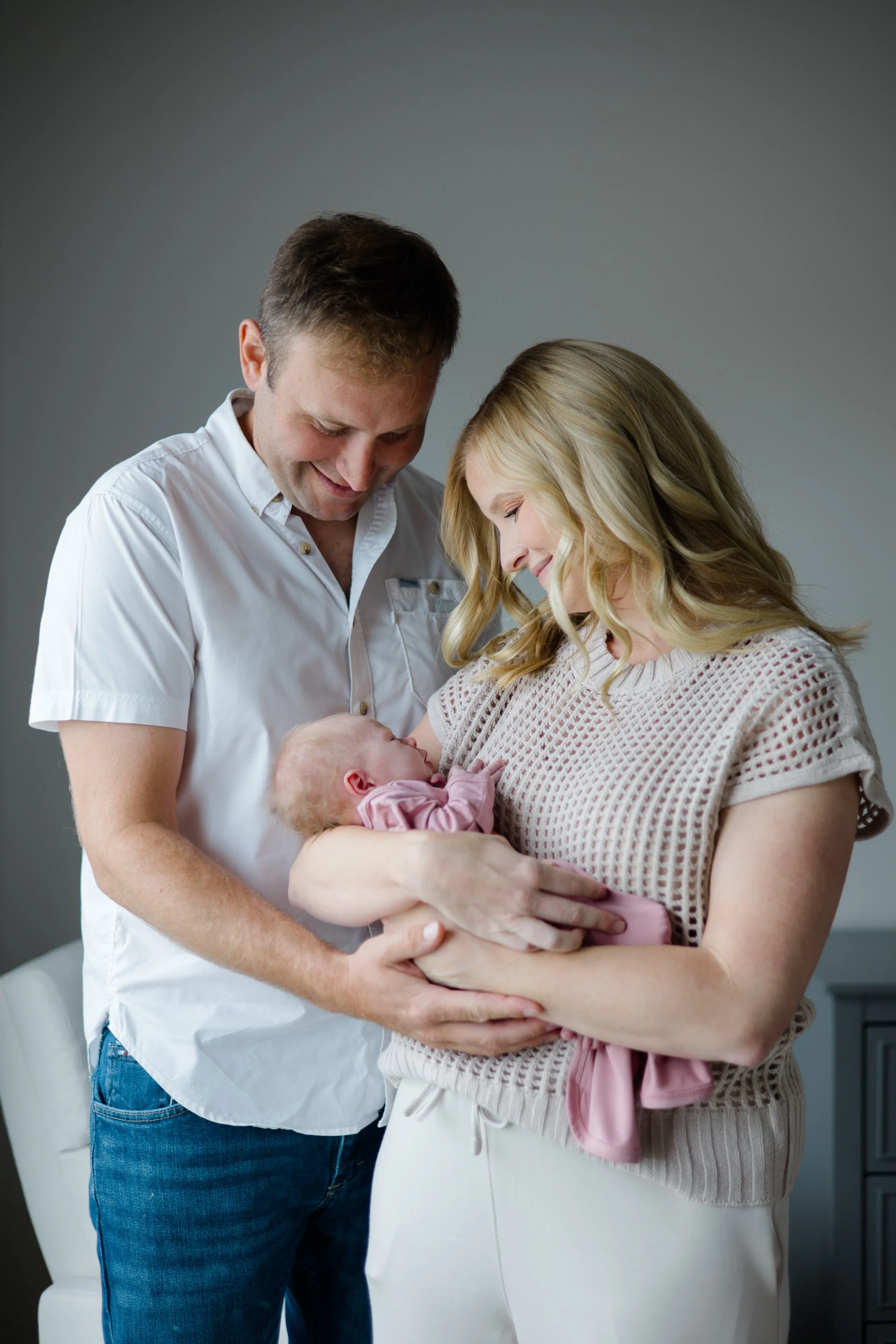 A woman holding a newborn baby, with a man standing beside them, all smiling and looking down at the baby.