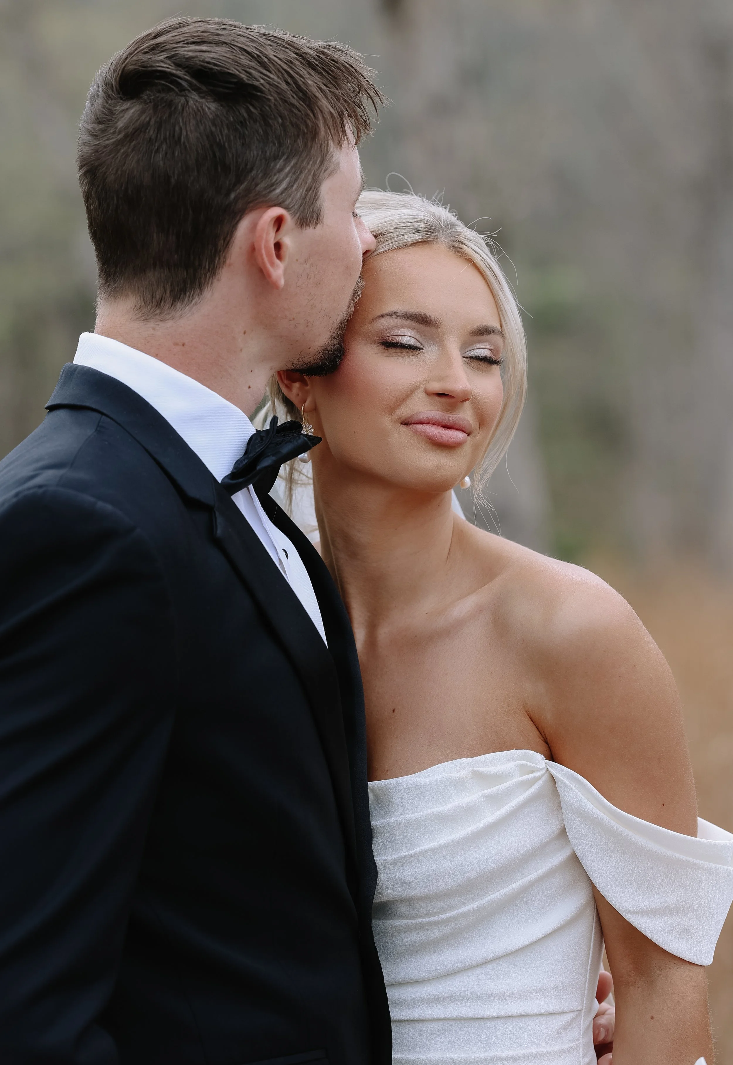 A man in a tuxedo and a woman in a white off-the-shoulder dress standing close together outdoors, with the man's face near the woman's temple and the woman's eyes closed, smiling gently.
