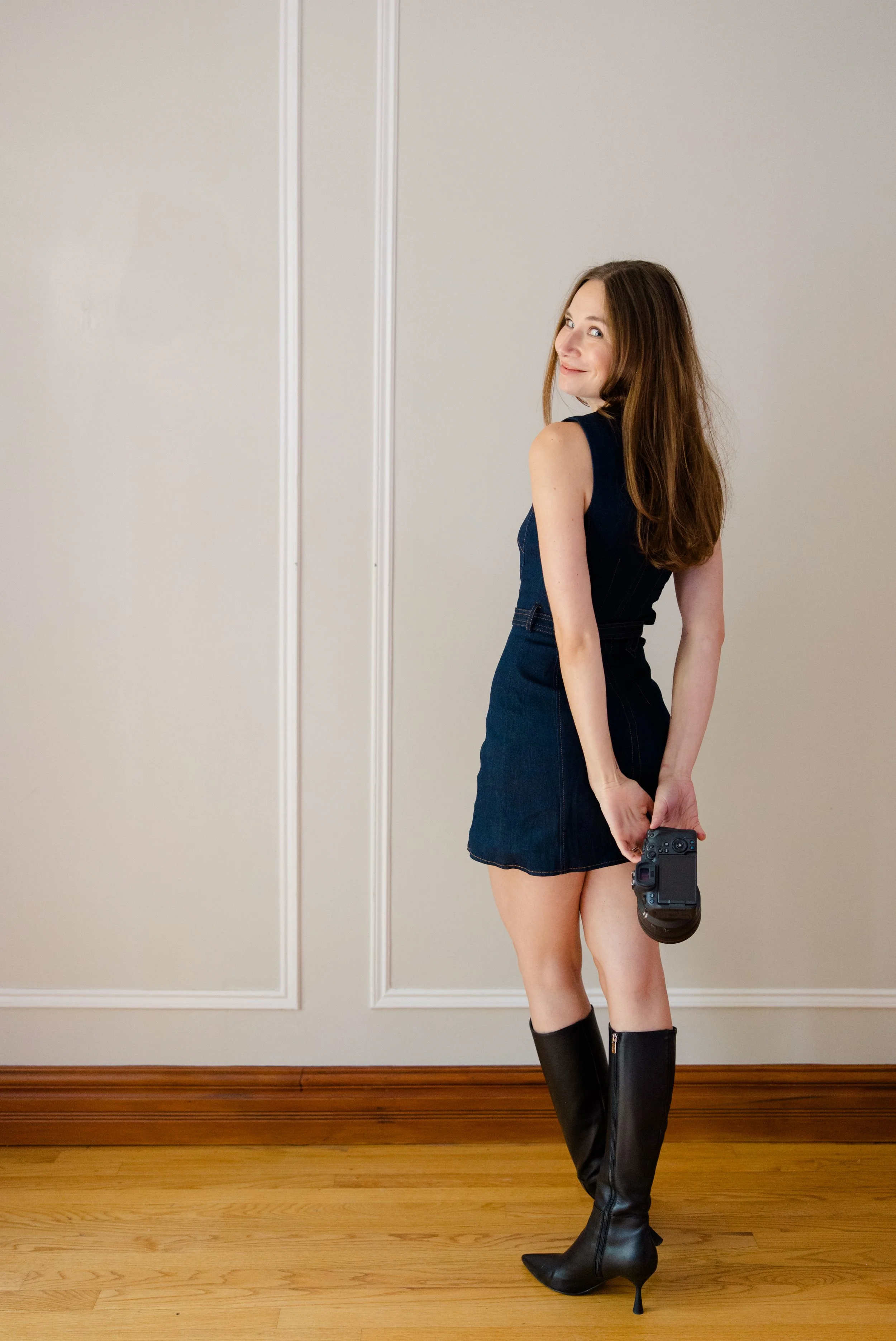 A woman standing in front of a beige wall with wooden trim, holding a camera in her right hand, wearing a sleeveless dark dress and high-heeled black boots.