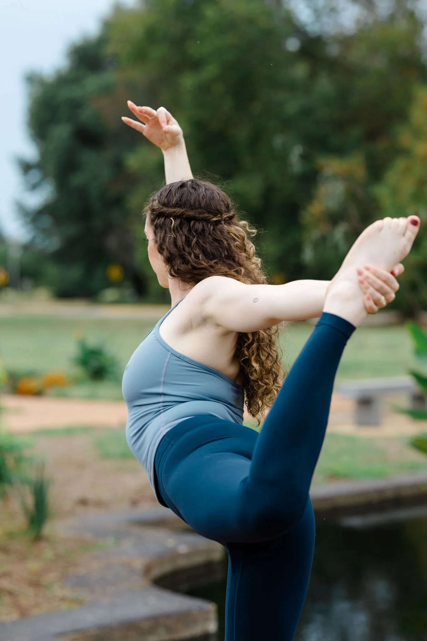 A woman with long curly hair practices yoga outdoors on a grassy area by a small stream. She is in a backbend pose with one arm raised and the other holding her foot.