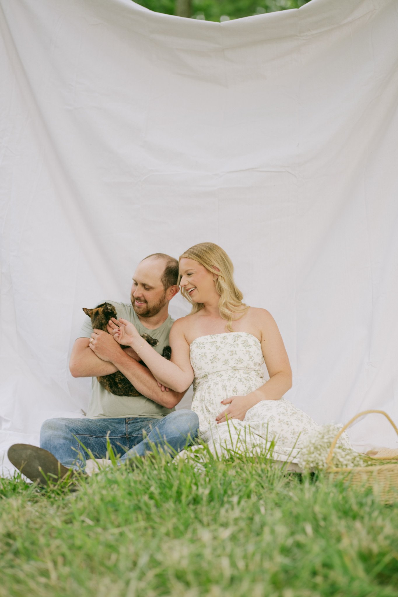 A couple sitting outdoors on green grass, with the man holding a small kitten, both smiling at the kitten, against a backdrop of a white sheet or fabric.