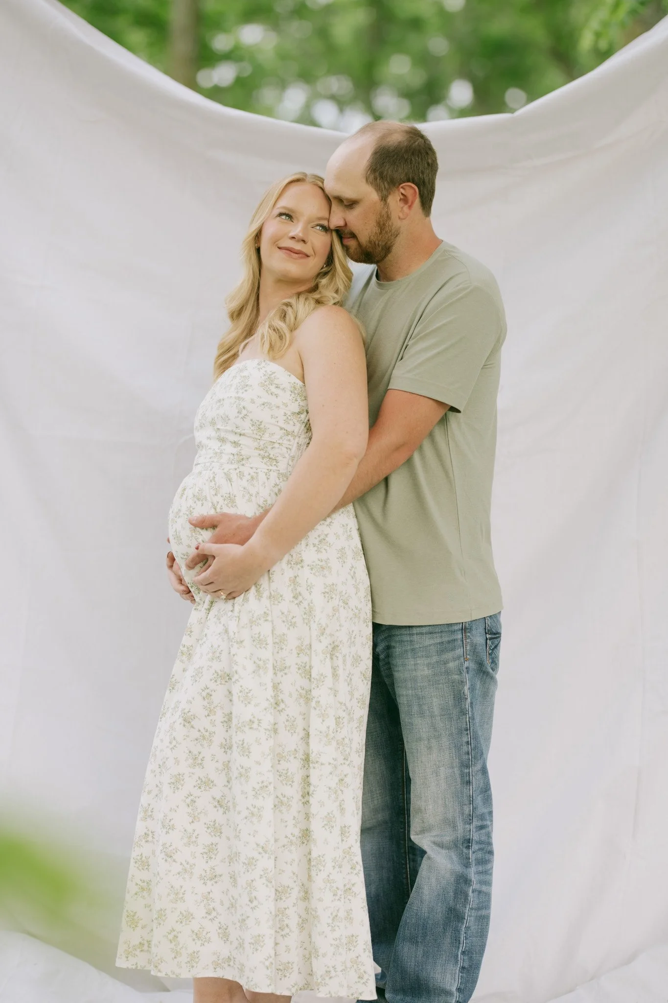 A pregnant woman in a floral dress and a man in jeans and a green t-shirt standing behind her, holding her belly and touching foreheads, outdoors with a white backdrop and green blurred trees in the background.