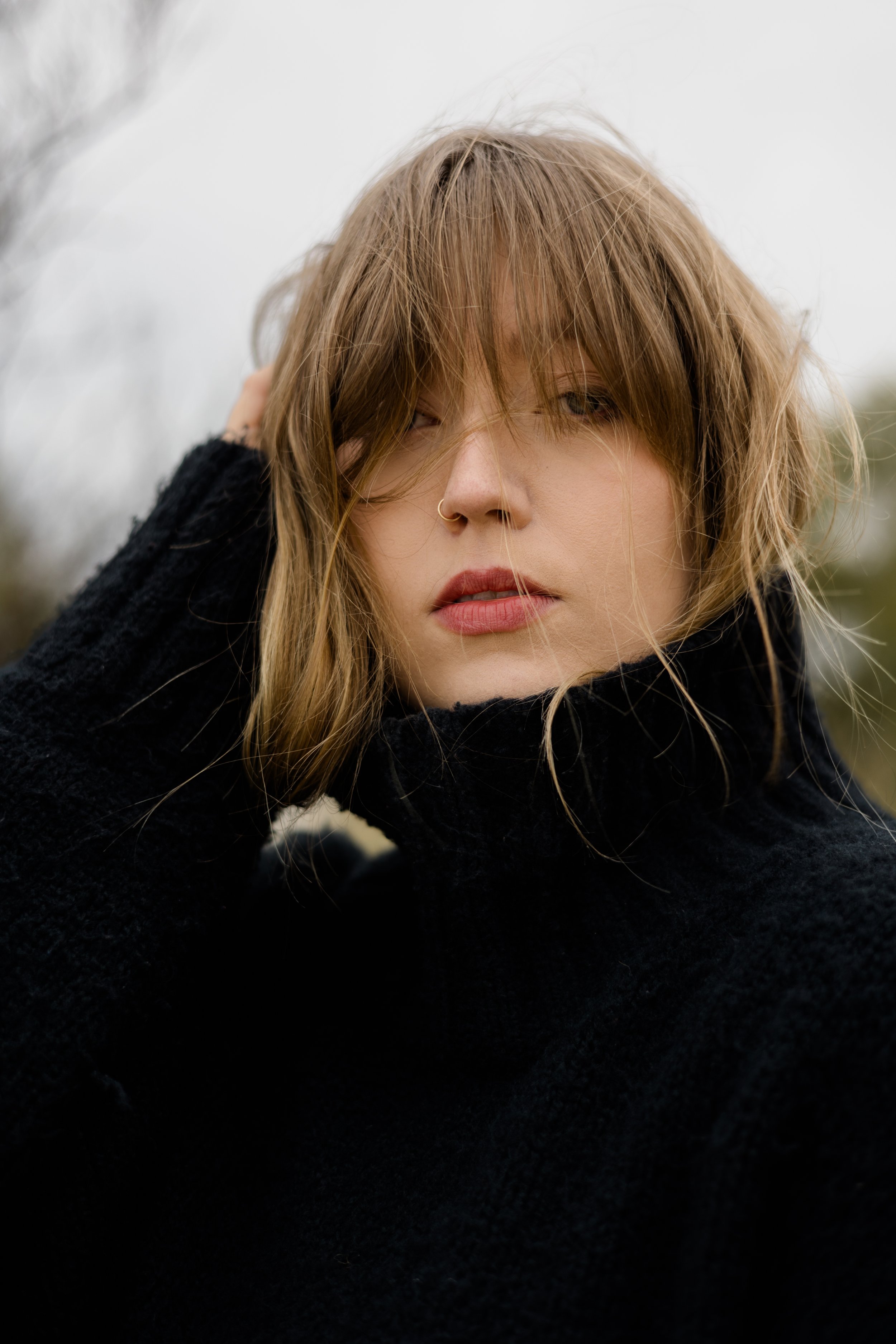 Close-up of a woman with light brown, slightly messy hair and a nose ring, wearing a black sweater, outdoors with a blurry background.