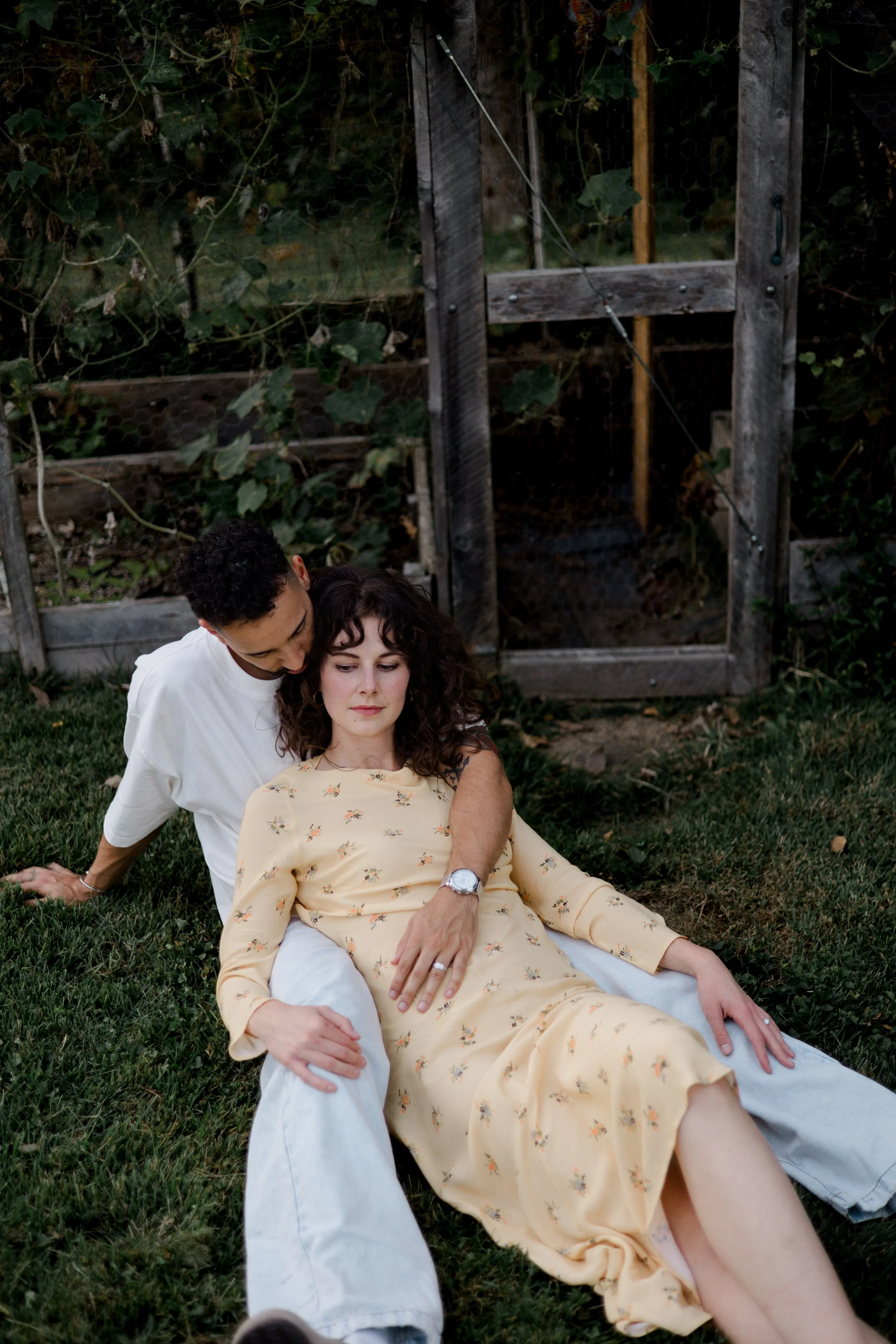 A young woman is lying on the grass outside with her eyes closed, wearing a yellow floral dress, and a man is sitting close beside her, placing his hand on her stomach while leaning close to her head. They are in a garden with a wooden and wire fence